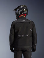 Giacca Alpinestars Cusco Drystar®, nero, vista posteriore di un pilota con casco nero e guanti, presenta ampi pannelli di ventilazione, logo bianco sul collo e marchio riflettente sulla parte inferiore della schiena, abbigliamento adventure touring