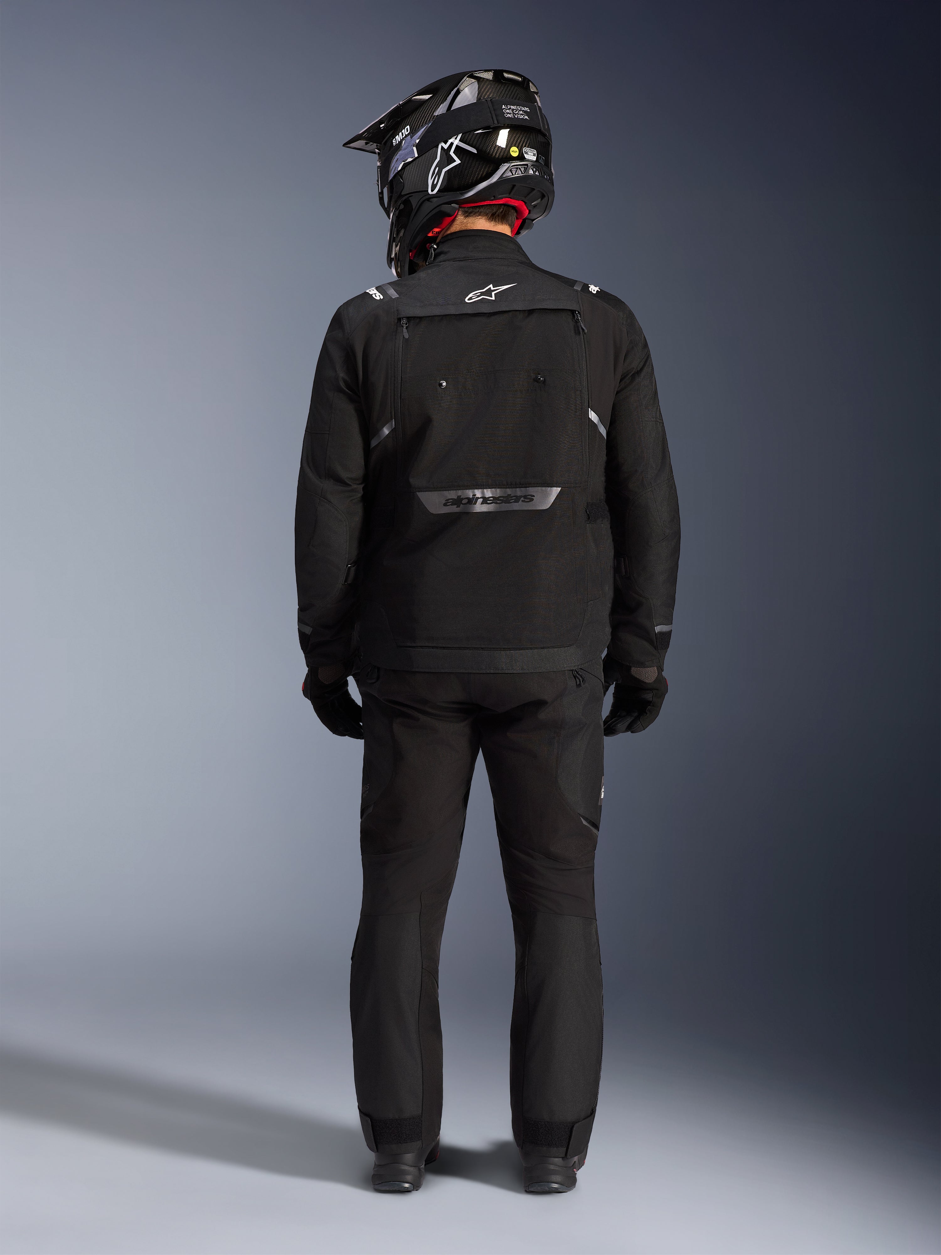 Alpinestars Giacca Cusco Drystar, giacca da moto adventure, nero, vista posteriore indossata con pantaloni coordinati e un casco bianco e nero, con marchio Alpinestars riflettente e ampi pannelli di ventilazione, robusta costruzione in tessuto per touring d'avventura su strada