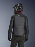 Pilota visto da dietro che indossa la giacca Alpinestars Cusco Drystar® in nero e grigio scuro, con un ampio pannello di ventilazione grigio sulla schiena e logo bianco, abbinata a un casco off-road nero e abbigliamento tecnico nero per l'adventure touring.