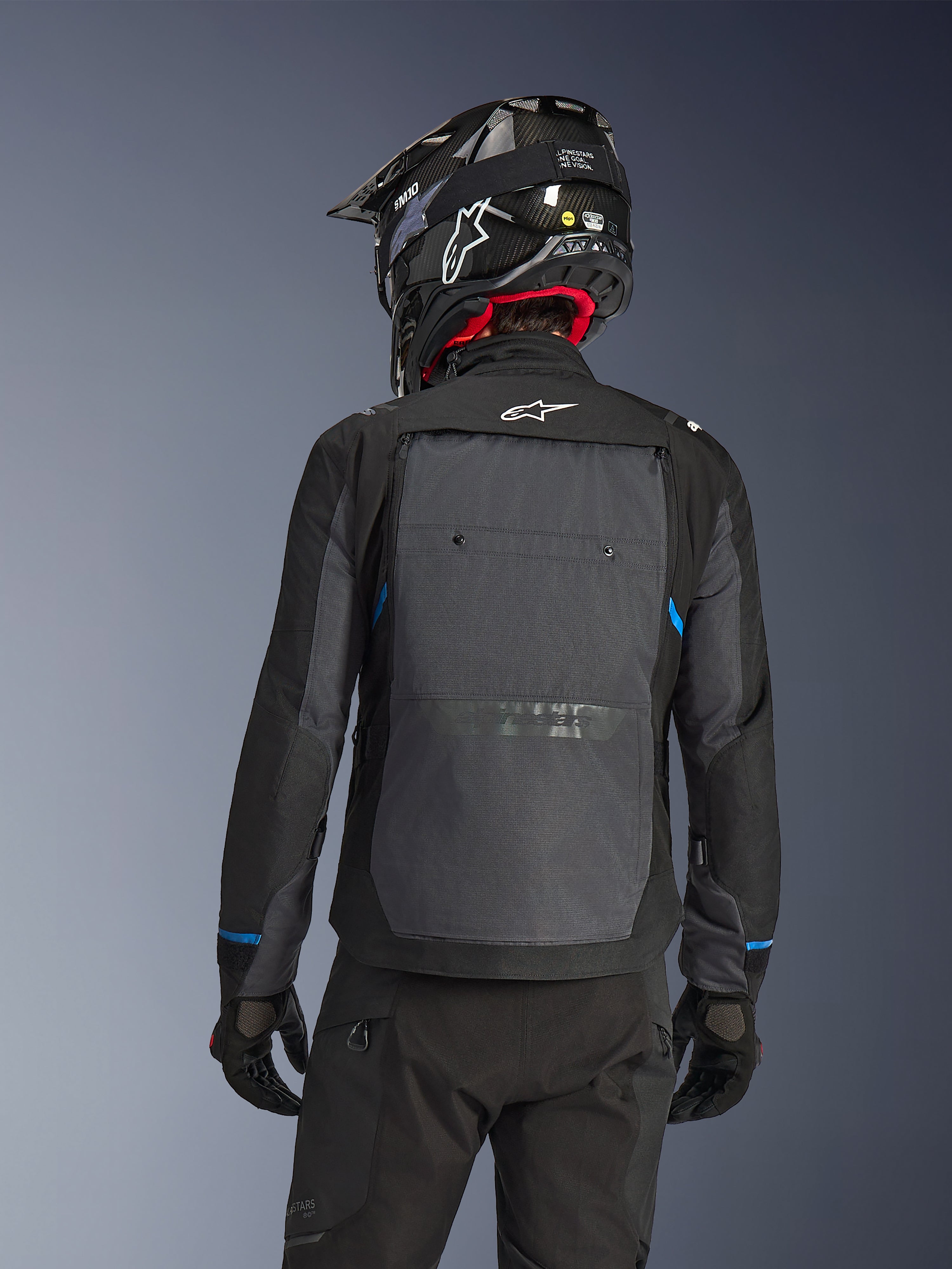 Pilota visto da dietro che indossa la giacca Alpinestars Cusco Drystar® in nero e grigio scuro, con un ampio pannello di ventilazione grigio sulla schiena e logo bianco, abbinata a un casco off-road nero e abbigliamento tecnico nero per l'adventure touring.
