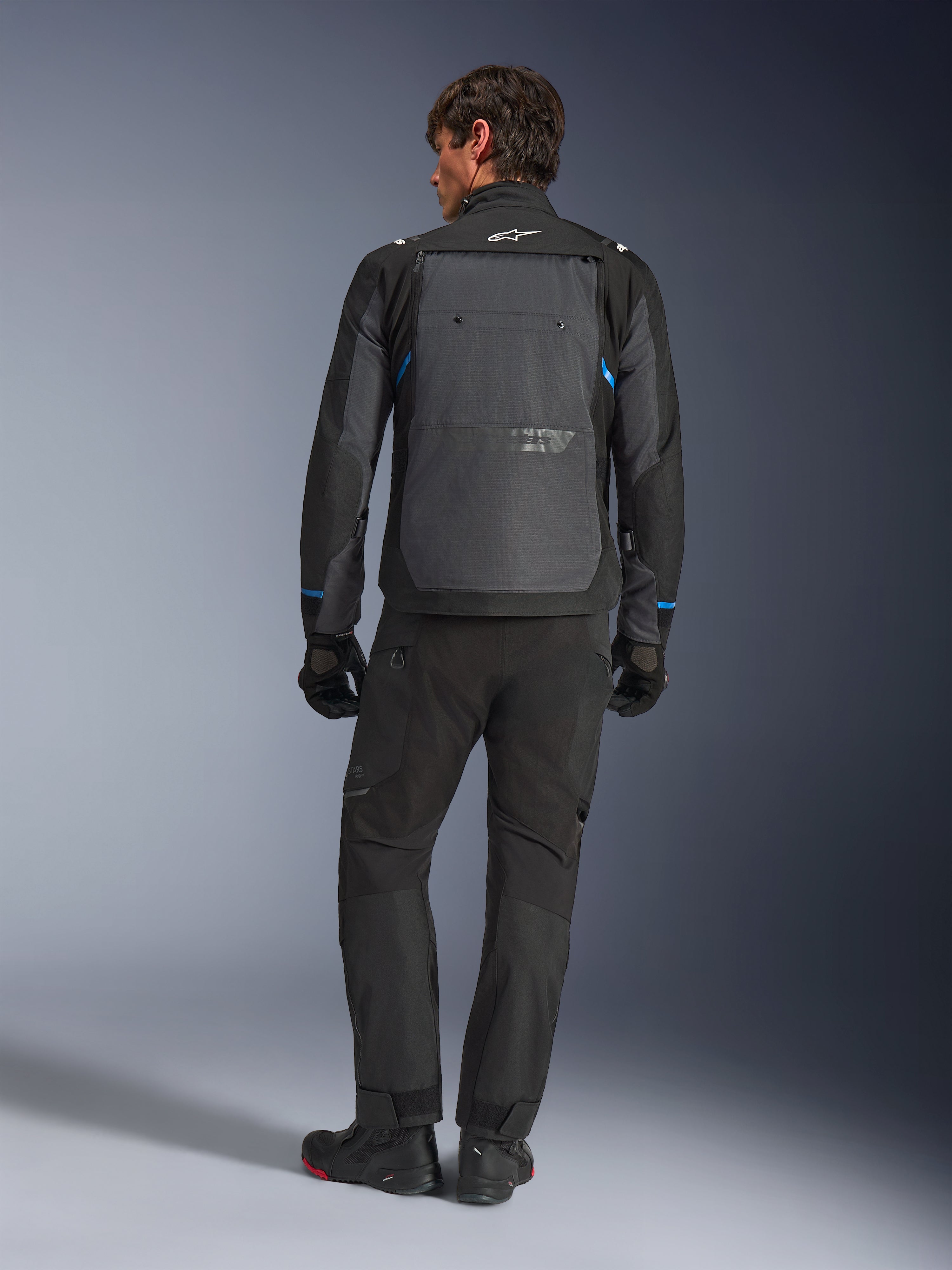 Giacca Alpinestars Cusco Drystar®, nero e grigio scuro con accenti blu, vista posteriore, indossata con pantaloni adventure e guanti neri abbinati, caratterizzata da un ampio pannello posteriore grigio e logo bianco sul colletto, progettata per il touring su strada e l'adventure touring