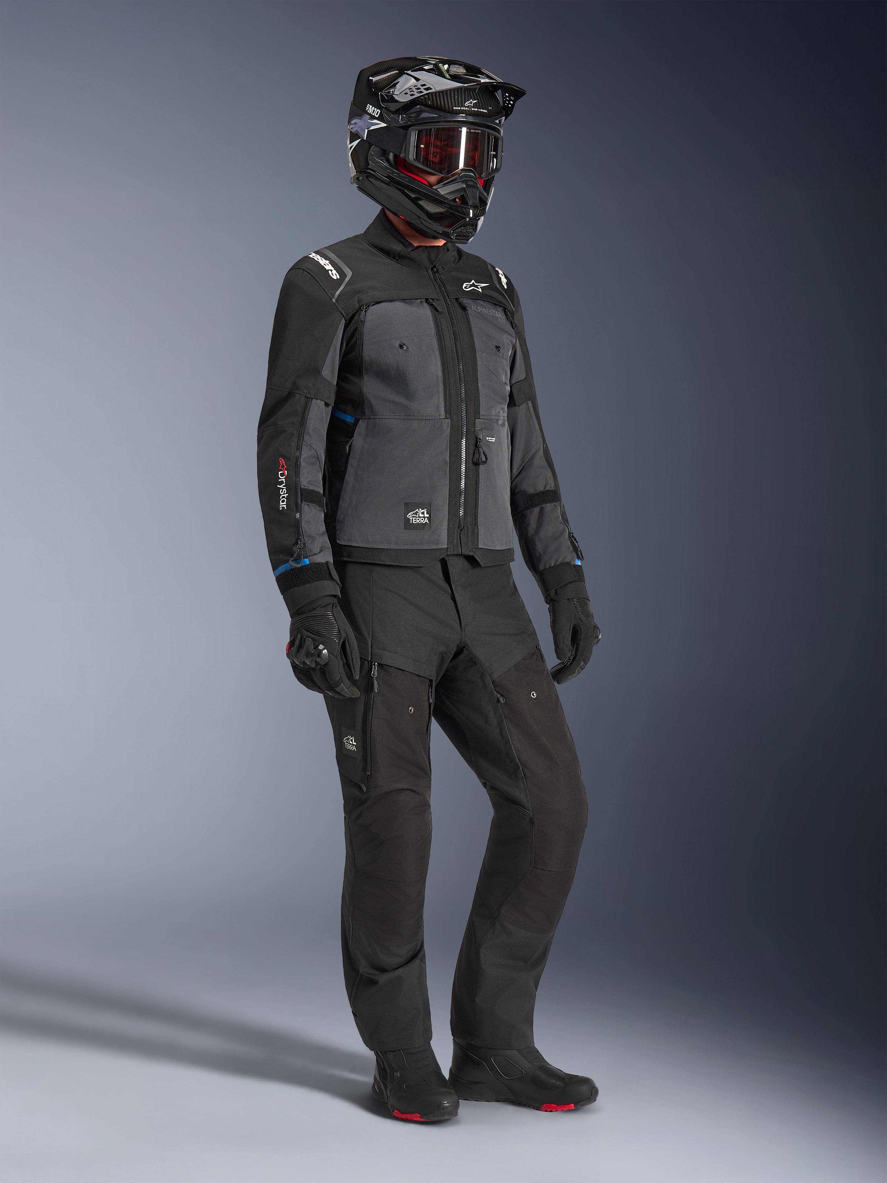 Modello che indossa la giacca Alpinestars Cusco Drystar® in Nero e Deep Gray, abbinata a un equipaggiamento da moto completo con casco nero, pantaloni tecnici coordinati, guanti e stivali, con costruzione in ripstop e softshell per il mototurismo stradale e adventure