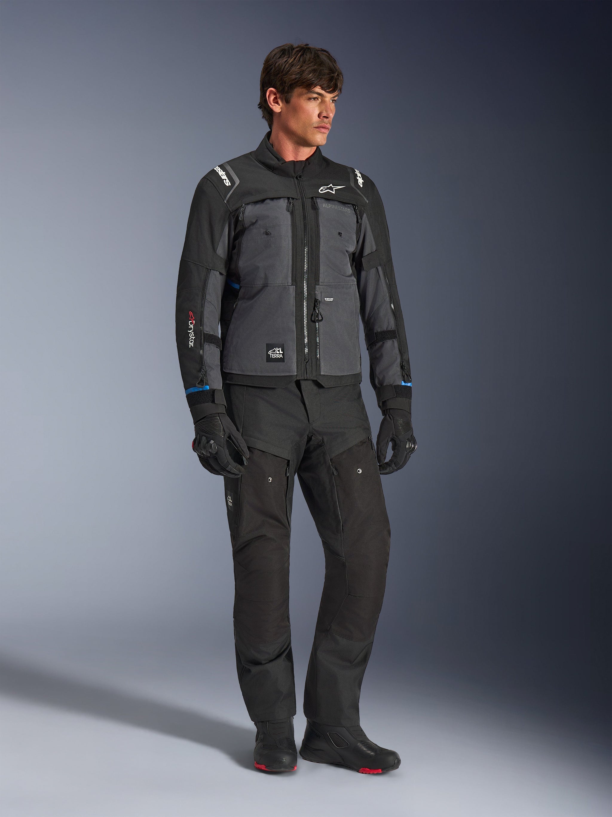 Giacca Alpinestars Cusco Drystar® in nero e grigio scuro, giacca tecnica da moto touring con pannelli multitono, indossata con pantaloni da moto e guanti neri coordinati, progettata per il touring su strada e adventure touring.