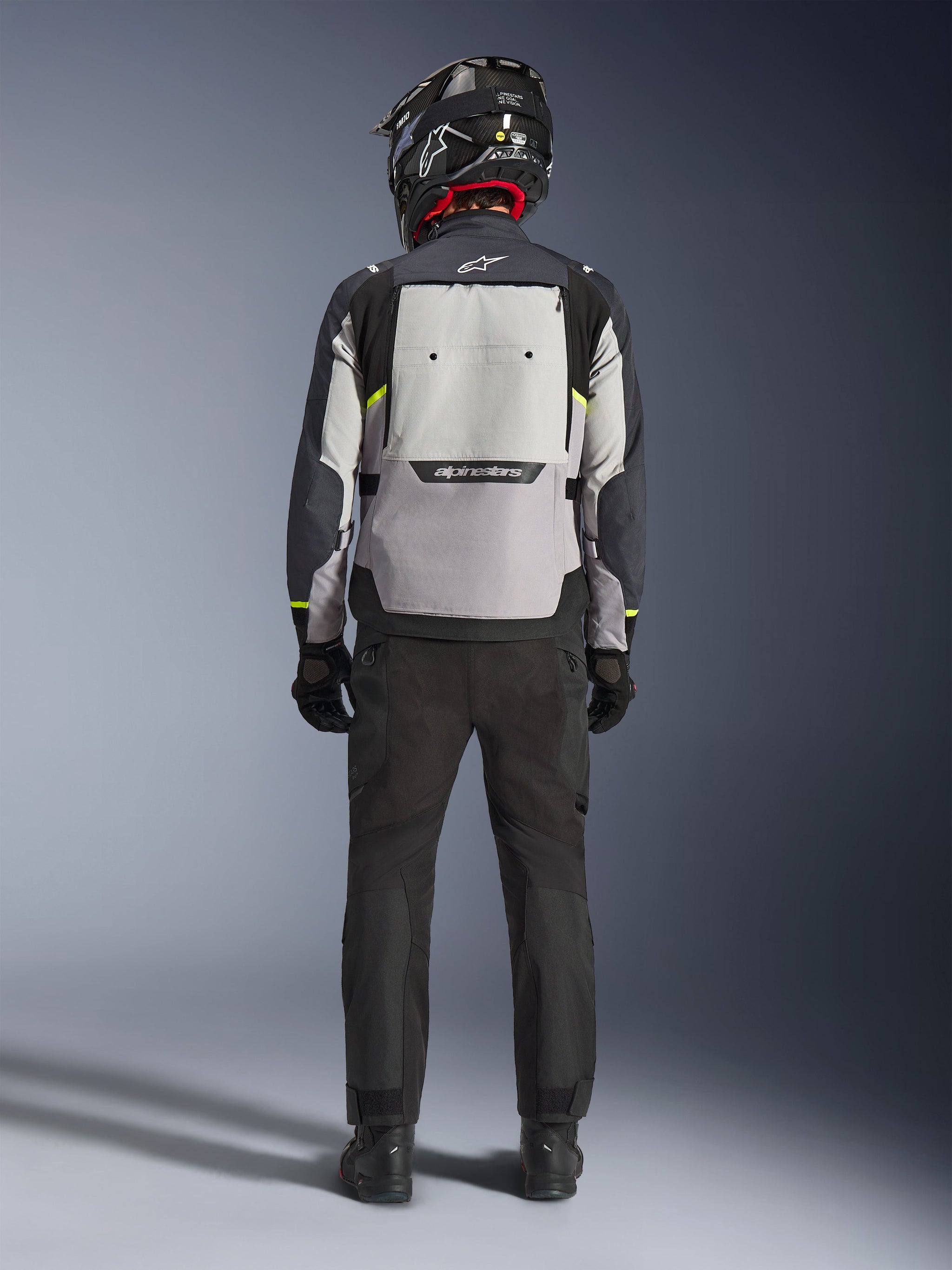 Motociclista visto da dietro che indossa la giacca Alpinestars Cusco Drystar® in Ice Gray, Mid Gray e Deep Gray, con un ampio pannello di ventilazione posteriore e logo Alpinestars, abbinata a pantaloni da moto neri e un casco nero, abbigliamento da moto adventure touring