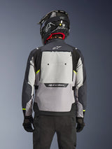 Giacca Alpinestars Cusco Drystar®, vista posteriore, Ice Gray, Mid Gray e Deep Gray, con ampio pannello di ventilazione posteriore e dettagli in giallo fluorescente ad alta visibilità, indossata con un casco adventure nero e pantaloni neri.