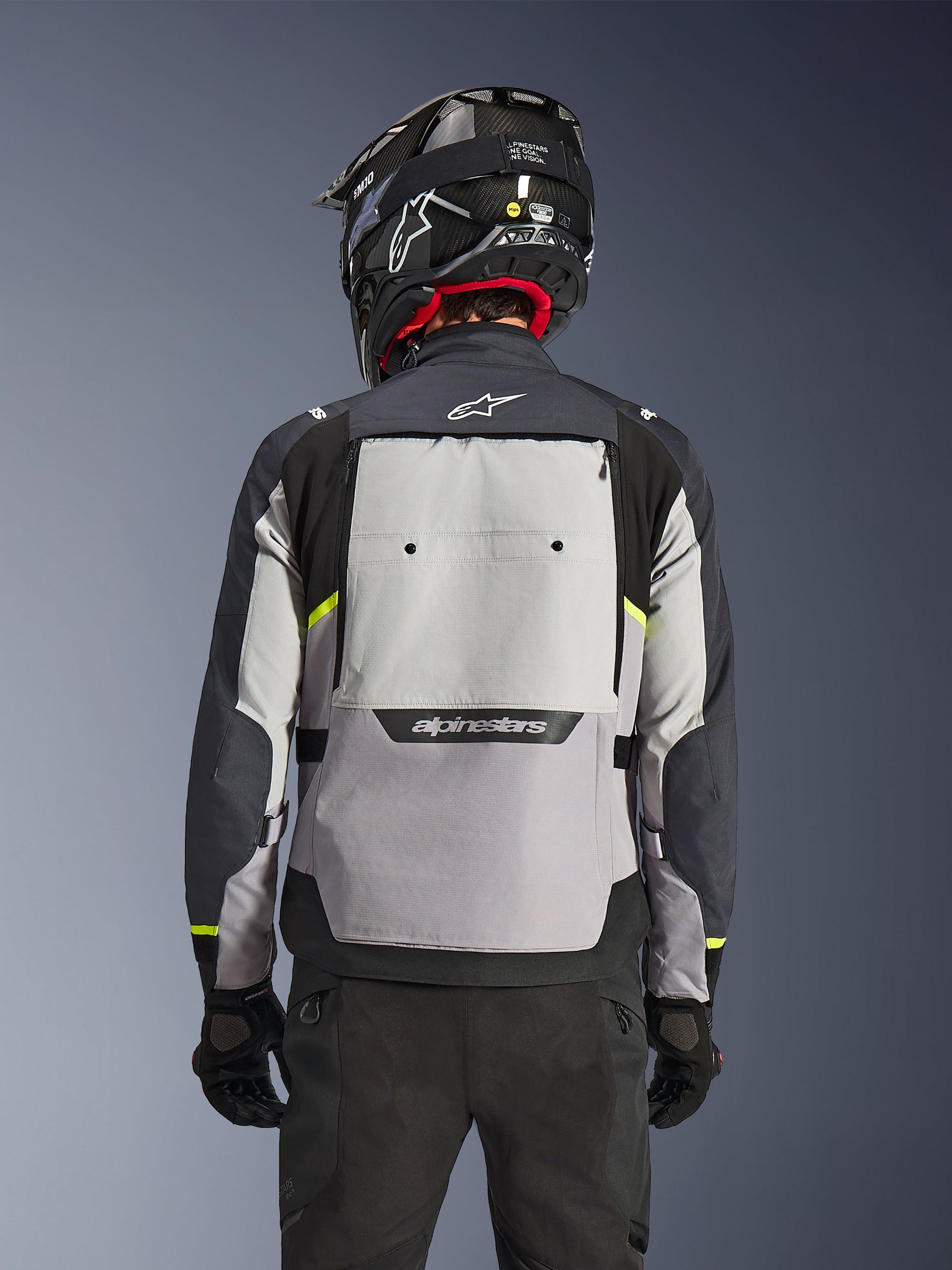Giacca Alpinestars Cusco Drystar®, vista posteriore, Ice Gray, Mid Gray e Deep Gray, con ampio pannello di ventilazione posteriore e dettagli in giallo fluorescente ad alta visibilità, indossata con un casco adventure nero e pantaloni neri.