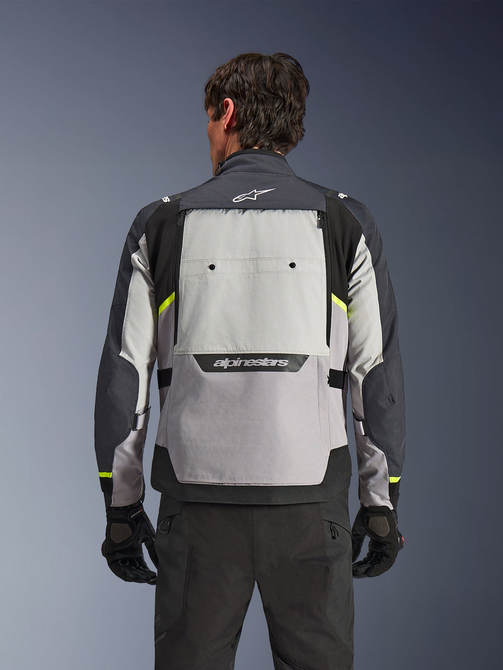 Vista posteriore di un pilota che indossa la giacca Alpinestars Cusco Drystar® in Ice Gray, Mid Gray e Deep Gray, con accenti giallo neon, un ampio pannello di ventilazione sulla schiena e logo Alpinestars, abbinata a pantaloni e guanti da moto neri.