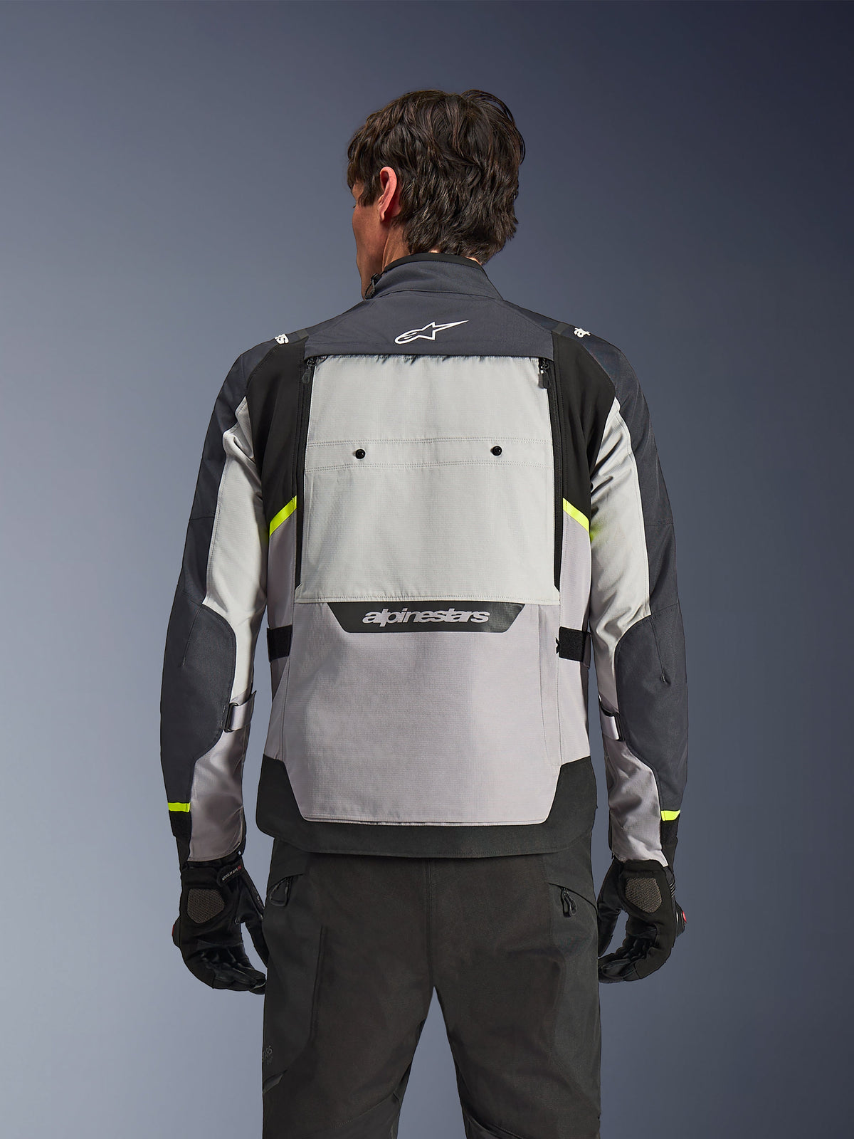 Vista posteriore di un pilota che indossa la giacca Alpinestars Cusco Drystar® in Ice Gray, Mid Gray e Deep Gray, con accenti giallo neon, un ampio pannello di ventilazione sulla schiena e logo Alpinestars, abbinata a pantaloni e guanti da moto neri.