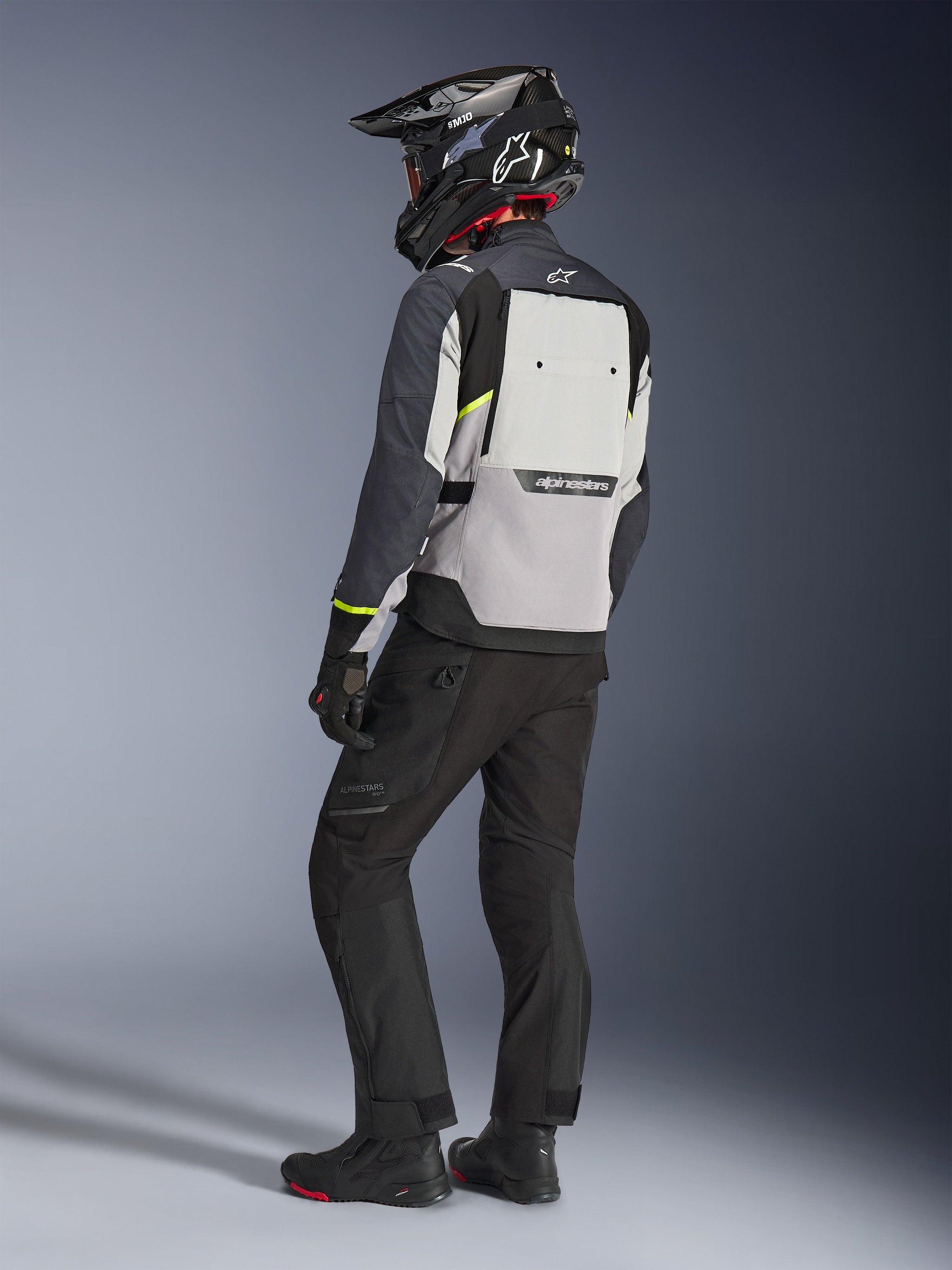 Giacca Alpinestars Cusco Drystar® in Ice Gray, Mid Gray e Deep Gray, vista posteriore su un pilota. Presenta accenti giallo fluorescente, ampio pannello di ventilazione posteriore con branding e costruzione in tessuto resistente. Indossata con pantaloni da moto neri e un casco adventure per un look touring completo.