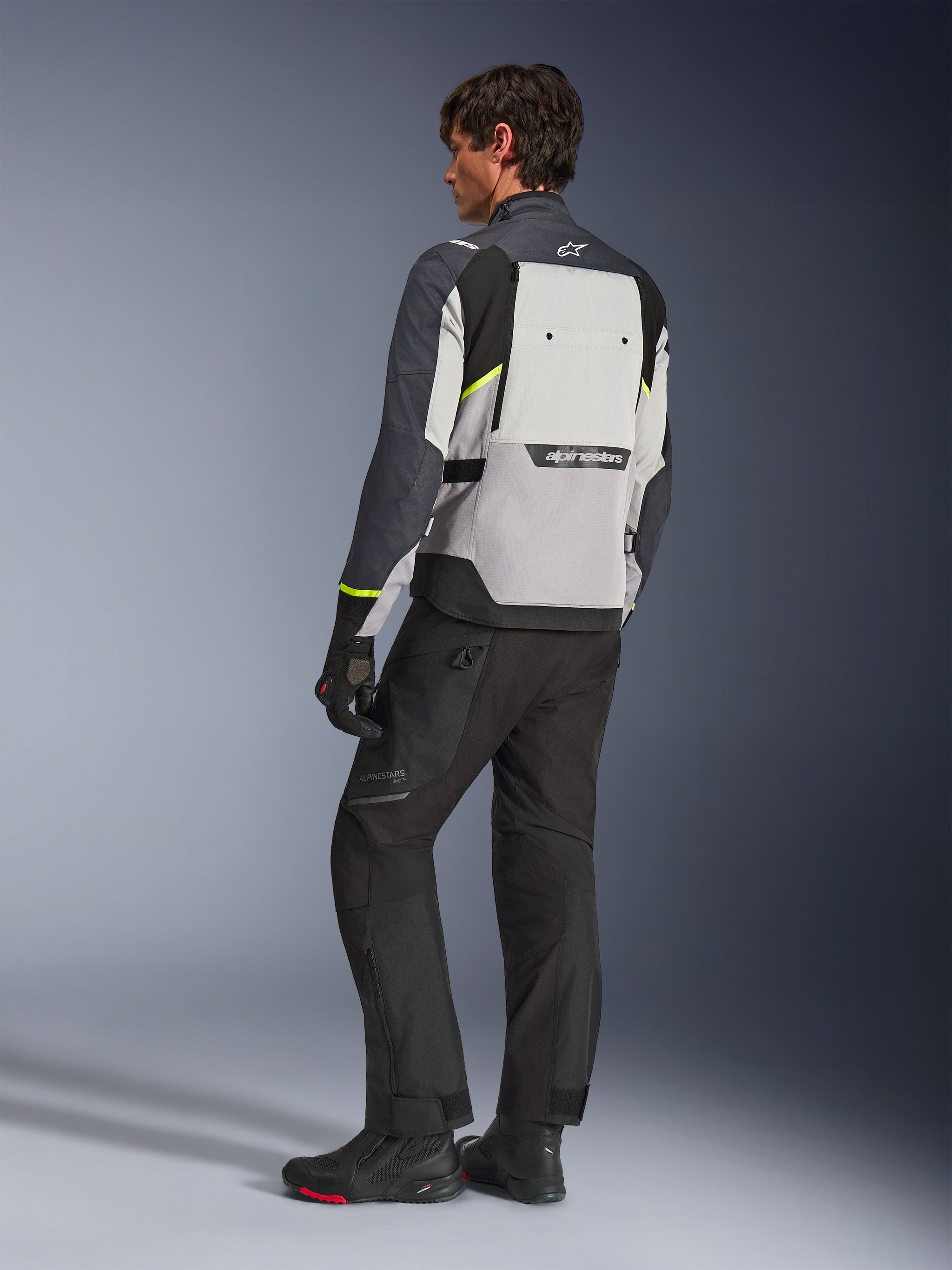 Giacca Alpinestars Cusco Drystar® in Ice Gray, Mid Gray e Deep Gray, vista posteriore su modello, giacca da adventure touring con accenti giallo neon, indossata con pantaloni da moto e stivali neri, con ampio pannello di ventilazione posteriore e logo Alpinestars.