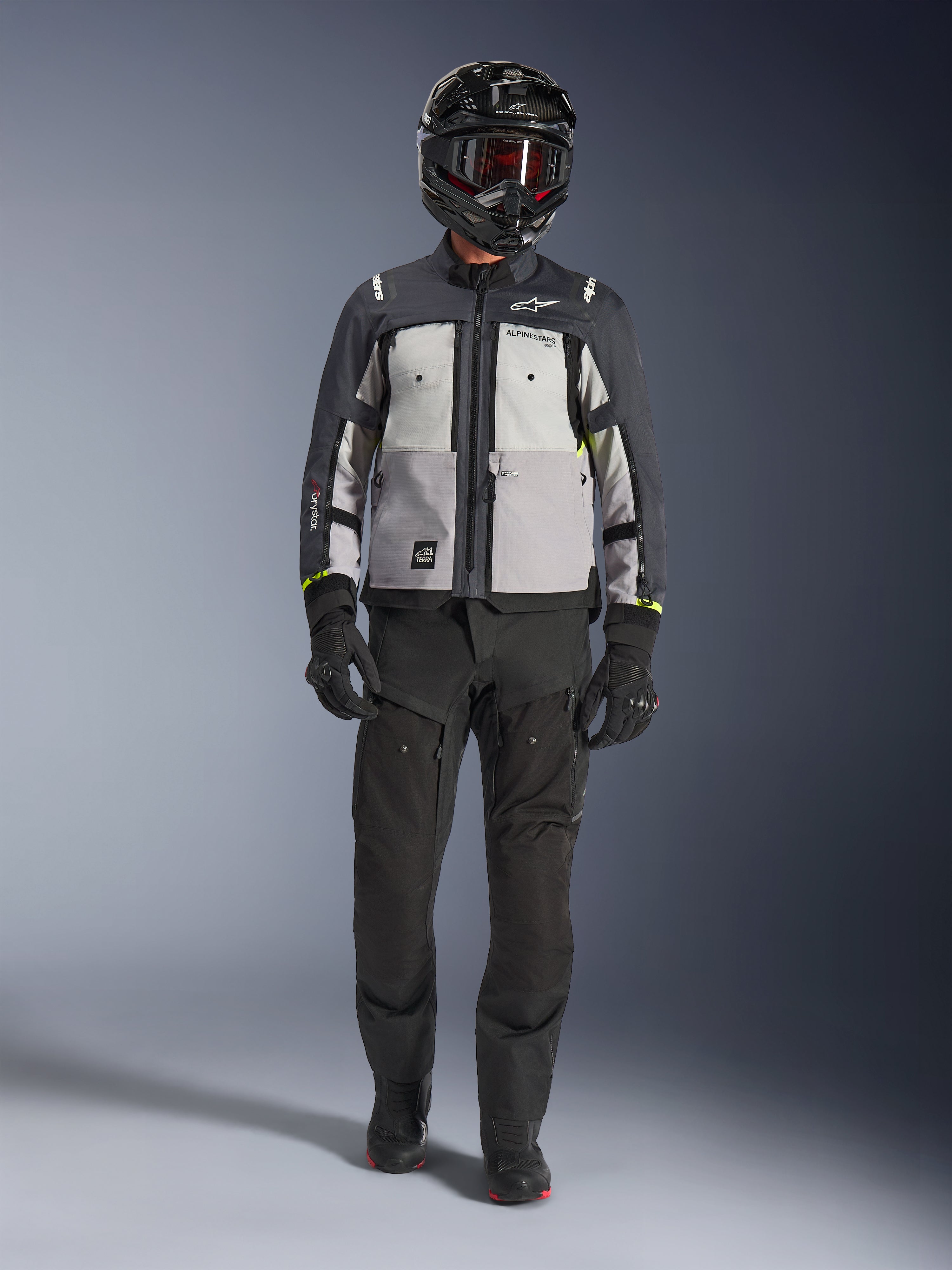 Giacca Alpinestars Cusco Drystar®, Ice Gray Mid Gray Deep Gray, giacca da moto adventure indossata con pantaloni da guida neri, guanti neri e casco nero con occhiali, caratterizzata da pannelli grigi color-block e dettagli tecnici per il touring su strada e adventure.