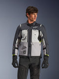 Giacca Alpinestars Cusco Drystar®, giacca da moto adventure touring, Ice Gray, Mid Gray e Deep Gray, indossata da un modello con pantaloni e guanti neri, caratterizzata da un design tecnico in tessuto a più pannelli, tasche sul petto, marchio 'Drystar' sulla manica e discreti accenti giallo neon sui polsini.