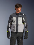 Giacca Alpinestars Cusco Drystar®, giacca da moto adventure touring, Ice Gray, Mid Gray e Deep Gray, indossata da un modello con pantaloni e guanti neri, caratterizzata da un design tecnico in tessuto a più pannelli, tasche sul petto, marchio 'Drystar' sulla manica e discreti accenti giallo neon sui polsini.
