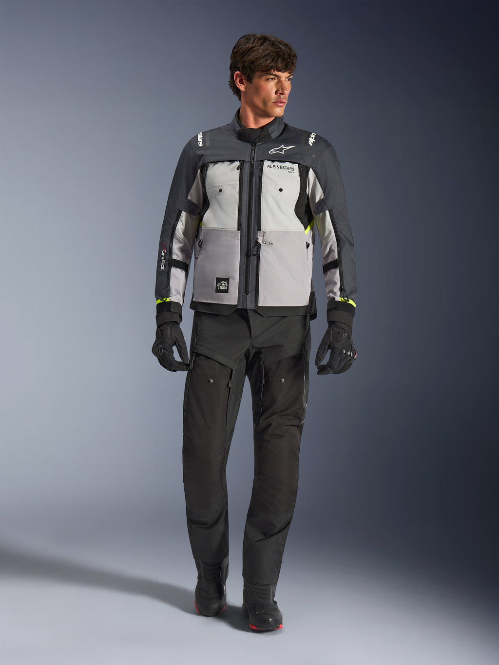 Giacca Alpinestars Cusco Drystar® in Ice Gray, Mid Gray e Deep Gray, indossata con pantaloni e guanti da moto adventure neri, giacca adventure touring in tessuto con pannelli grigi multi-tonali e accenti giallo neon, foto in studio con modello