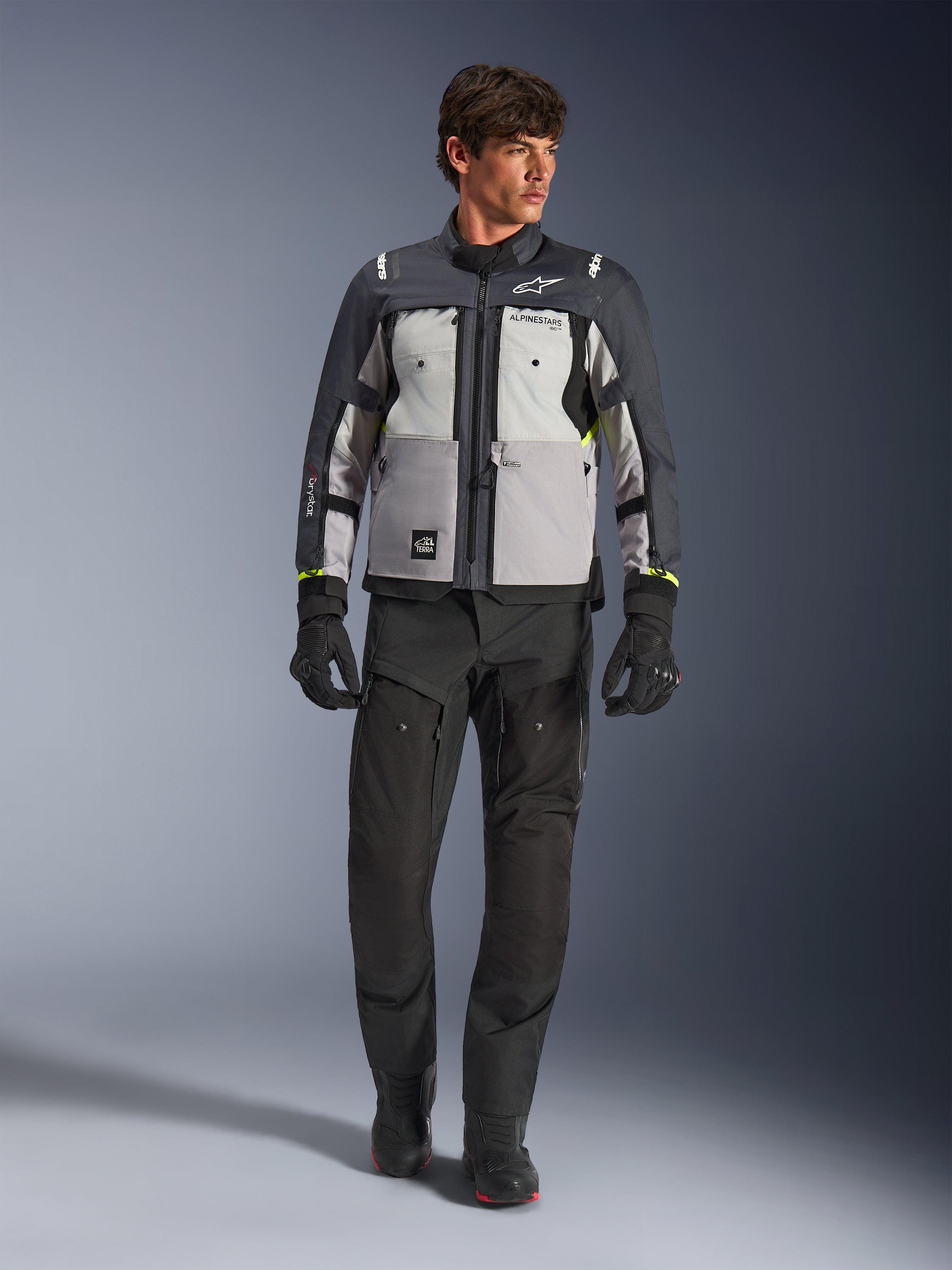 Giacca Alpinestars Cusco Drystar® in Ice Gray, Mid Gray e Deep Gray, indossata con pantaloni e guanti da moto adventure neri, giacca adventure touring in tessuto con pannelli grigi multi-tonali e accenti giallo neon, foto in studio con modello