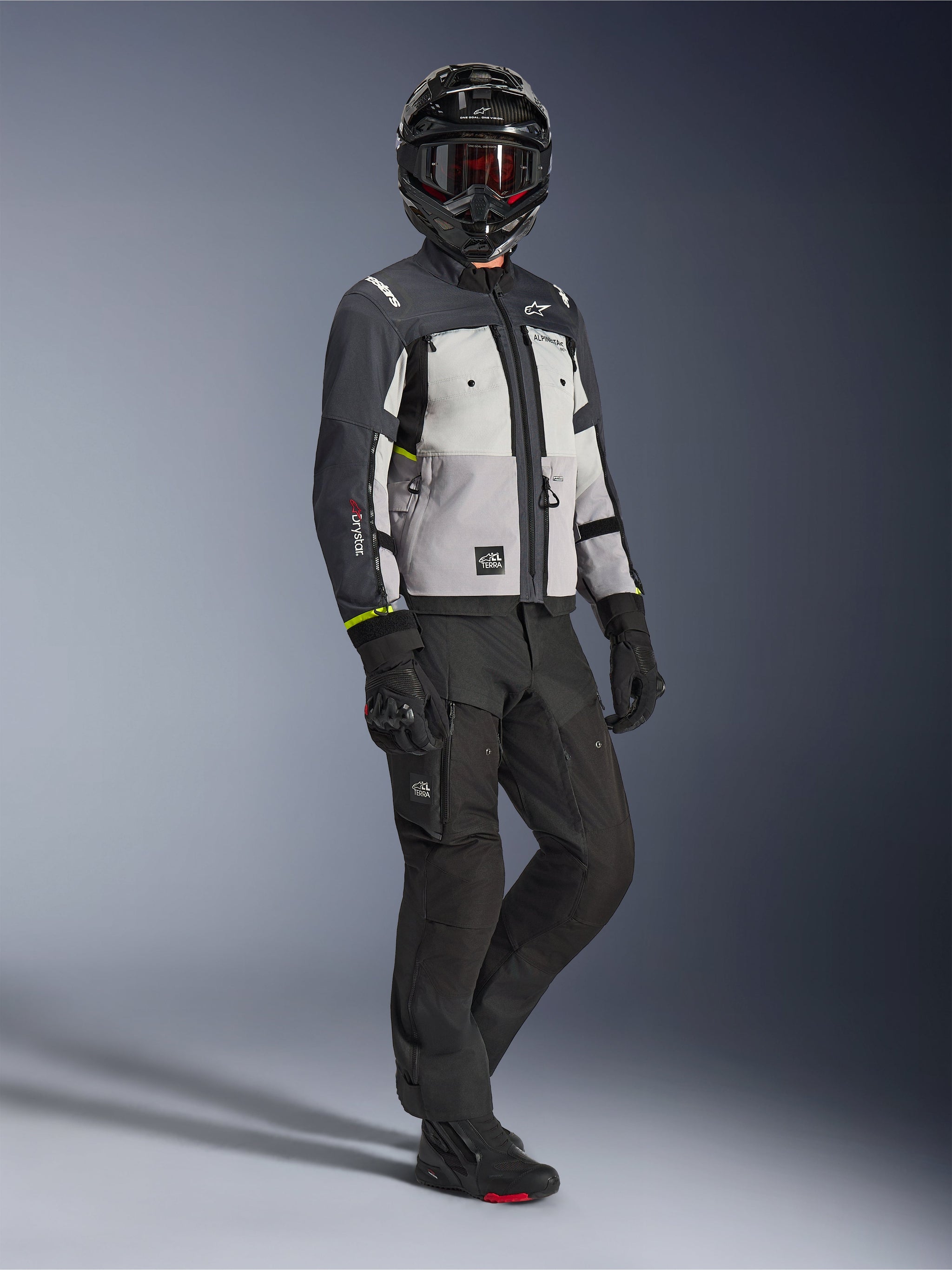 Giacca Alpinestars Cusco Drystar® in Ice Gray Mid Gray Deep Gray, inquadratura a figura intera di un pilota con equipaggiamento adventure completo, casco nero con maschera rossa, pantaloni e guanti neri abbinati, giacca da moto in tessuto con pannelli di ventilazione e membrana impermeabile