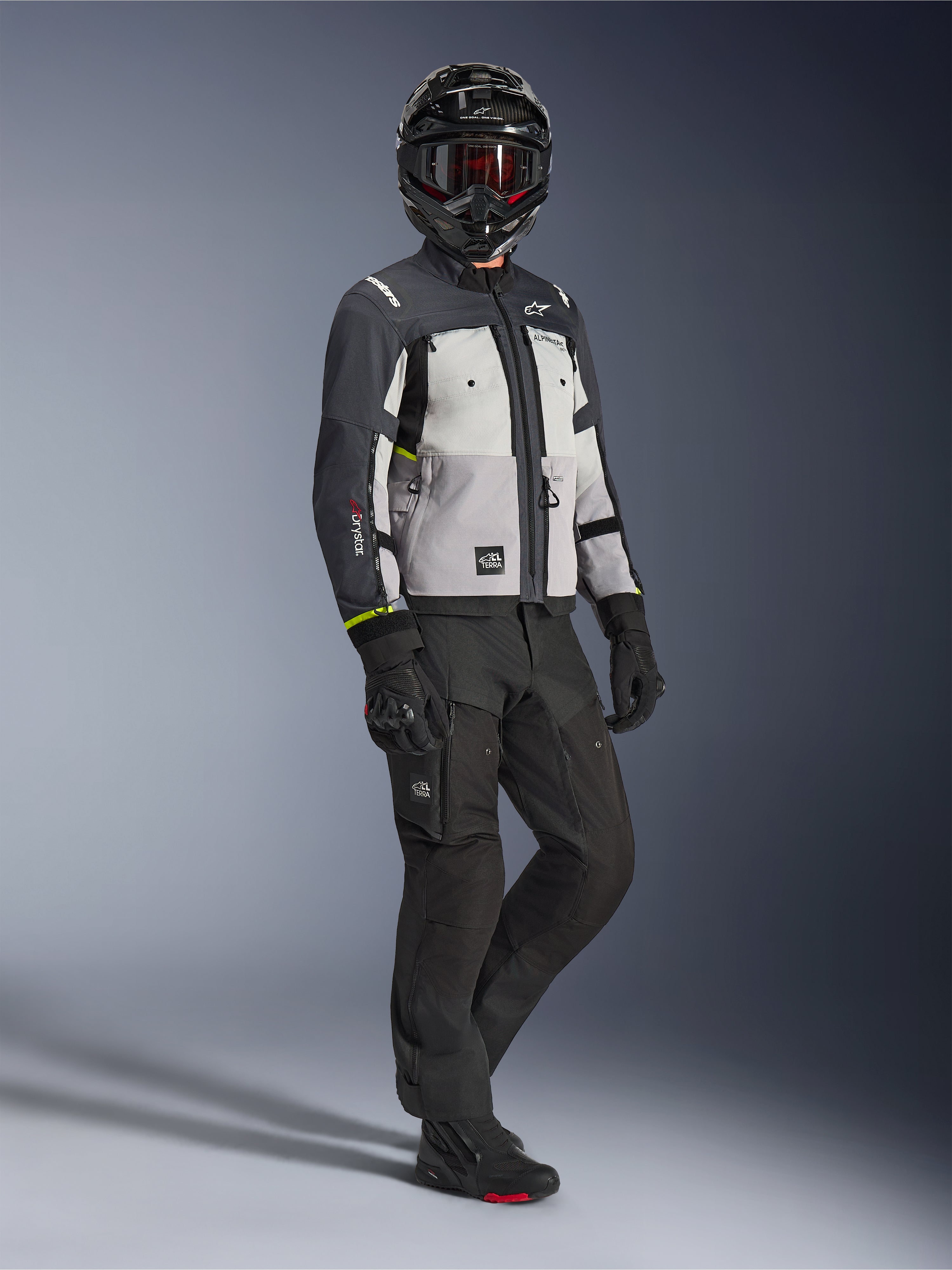 Giacca Alpinestars Cusco Drystar® in Ice Gray Mid Gray Deep Gray, inquadratura a figura intera di un pilota con equipaggiamento adventure completo, casco nero con maschera rossa, pantaloni e guanti neri abbinati, giacca da moto in tessuto con pannelli di ventilazione e membrana impermeabile