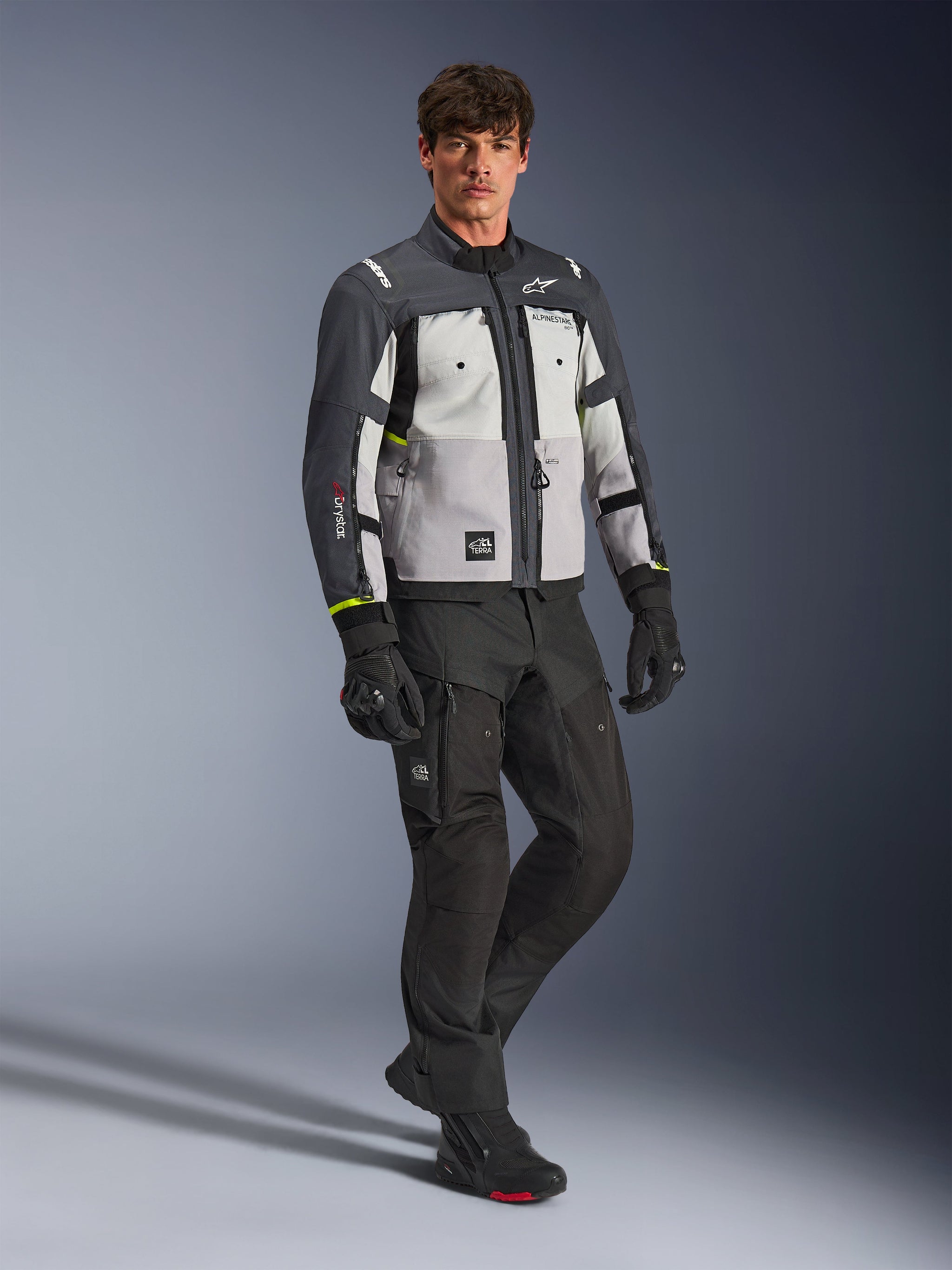 Giacca Alpinestars Cusco Drystar®, giacca da moto adventure, colori Ice Gray, Mid Gray e Deep Gray, indossata da un modello con pantaloni e guanti neri, pannelli grigi multi-tono in tessuto resistente, con prese d'aria con zip e logo Alpinestars, fotografia professionale in studio per abbigliamento da strada e adventure.