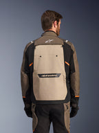Giacca Alpinestars Cusco Drystar®, Walnut Aluminum Black, vista posteriore, ampio pannello posteriore color sabbia con logo nero, accenti arancioni, indossata con pantaloni e guanti coordinati, progettata per l'adventure touring.