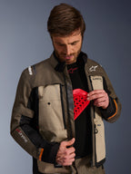 Giacca Alpinestars Cusco Drystar®, Walnut Aluminum Black, indossata da un uomo che mostra la tasca per la protezione del torace con un inserto protettivo rosso, giacca da moto in tessuto con prese d'aria con cerniera e loghi per adventure touring.