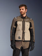 Giacca Alpinestars Cusco Drystar®, giacca da moto adventure, Walnut Aluminum Black, indossata da un modello con guanti neri, con pannelli sul petto color sabbia e maniche nere con marchio Alpinestars e toppa All Terra, abbigliamento protettivo da touring per ogni condizione atmosferica