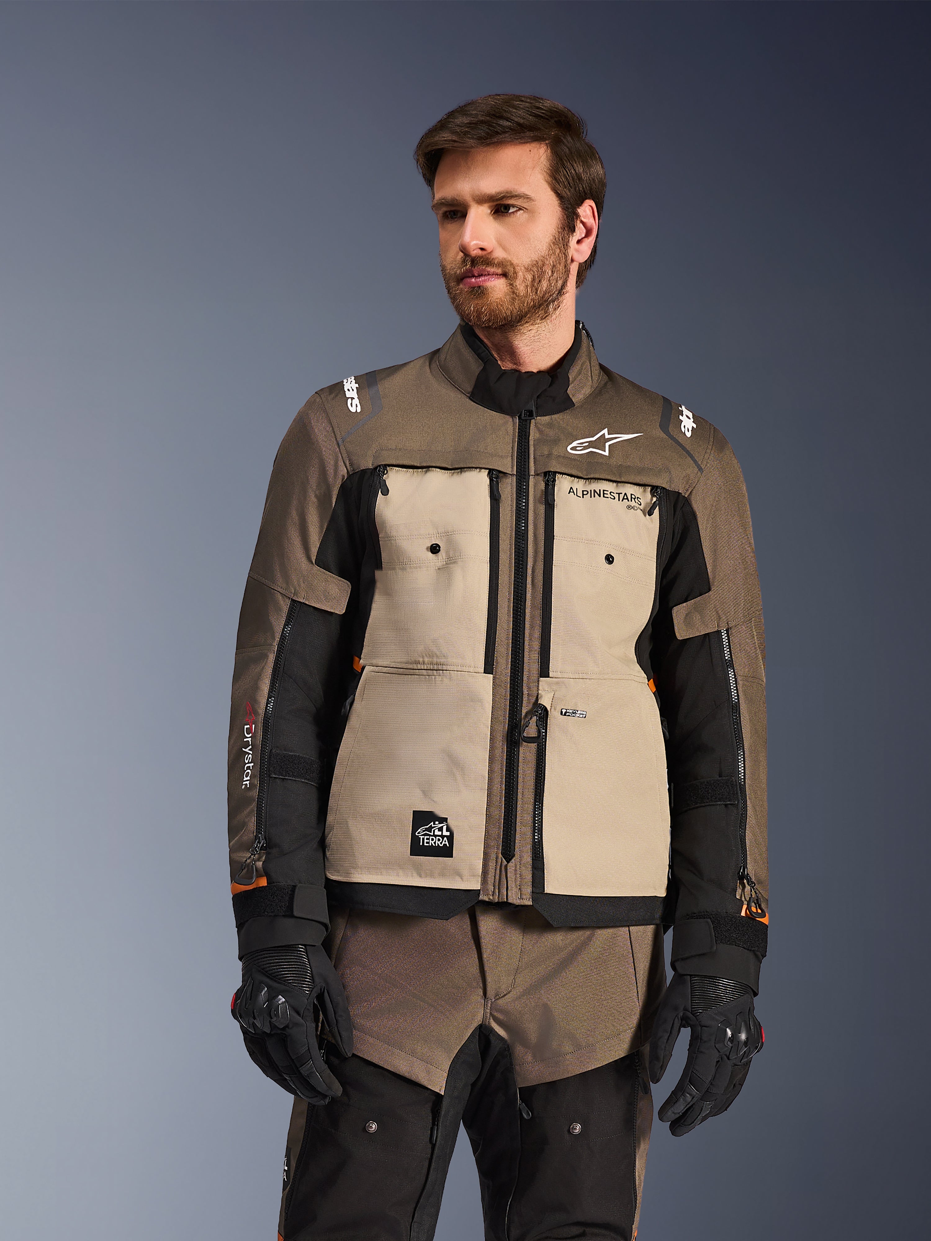 Giacca Alpinestars Cusco Drystar®, giacca da moto adventure, Walnut Aluminum Black, indossata da un modello con guanti neri, con pannelli sul petto color sabbia e maniche nere con marchio Alpinestars e toppa All Terra, abbigliamento protettivo da touring per ogni condizione atmosferica