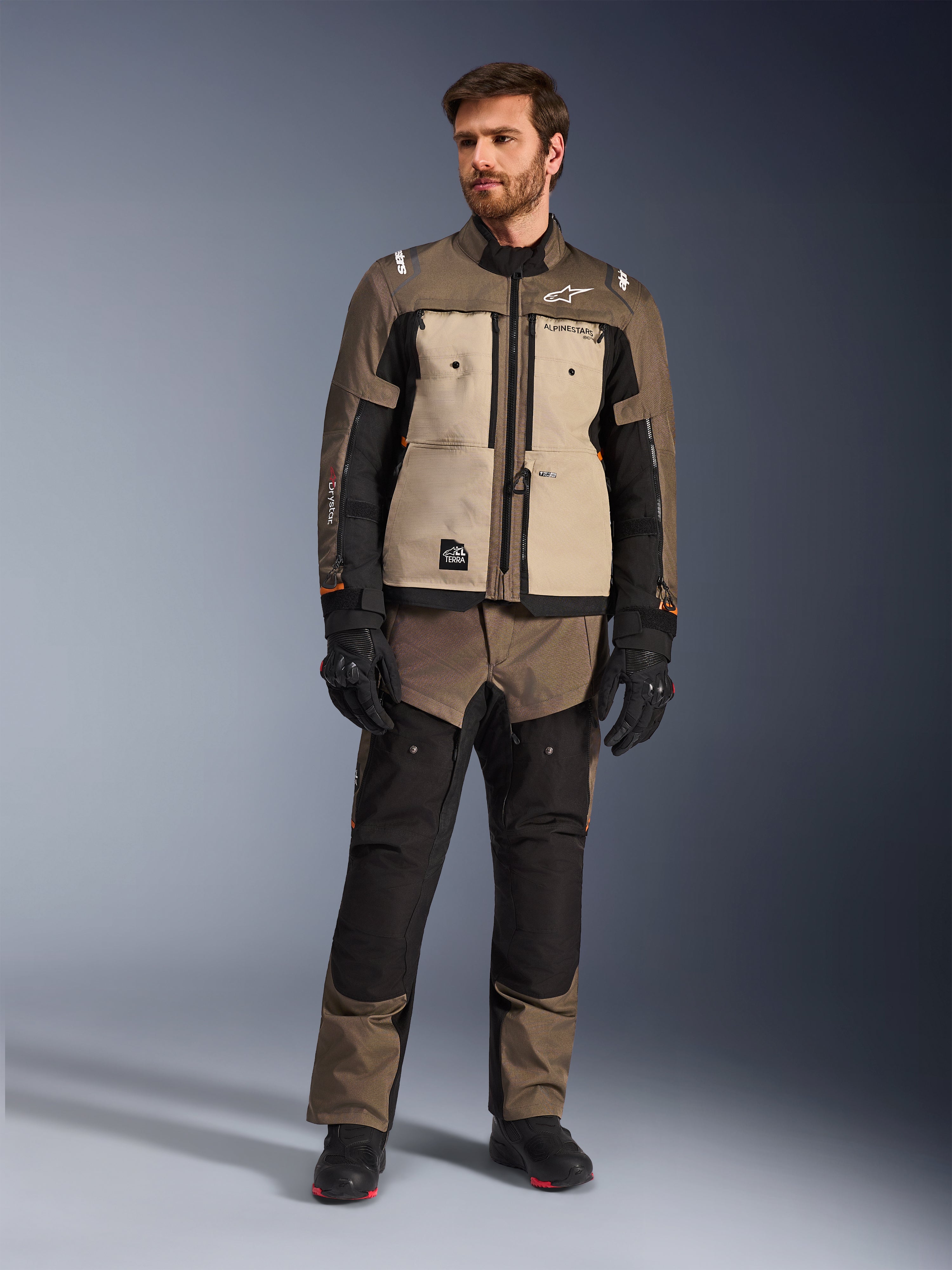 Giacca Alpinestars Cusco Drystar® in Walnut Aluminum Black, indossata da un motociclista con pantaloni adventure touring abbinati, guanti e stivali neri, con pannelli sul petto color sabbia, maniche nere con sottili accenti arancioni e cerniere di ventilazione funzionali per la guida su strada.