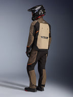 Giacca Alpinestars Cusco Drystar®, giacca adventure touring, Walnut Aluminum Black, vista posteriore che mostra il pannello posteriore marrone chiaro e i dettagli arancioni, indossata con pantaloni coordinati e casco nero in fibra di carbonio.