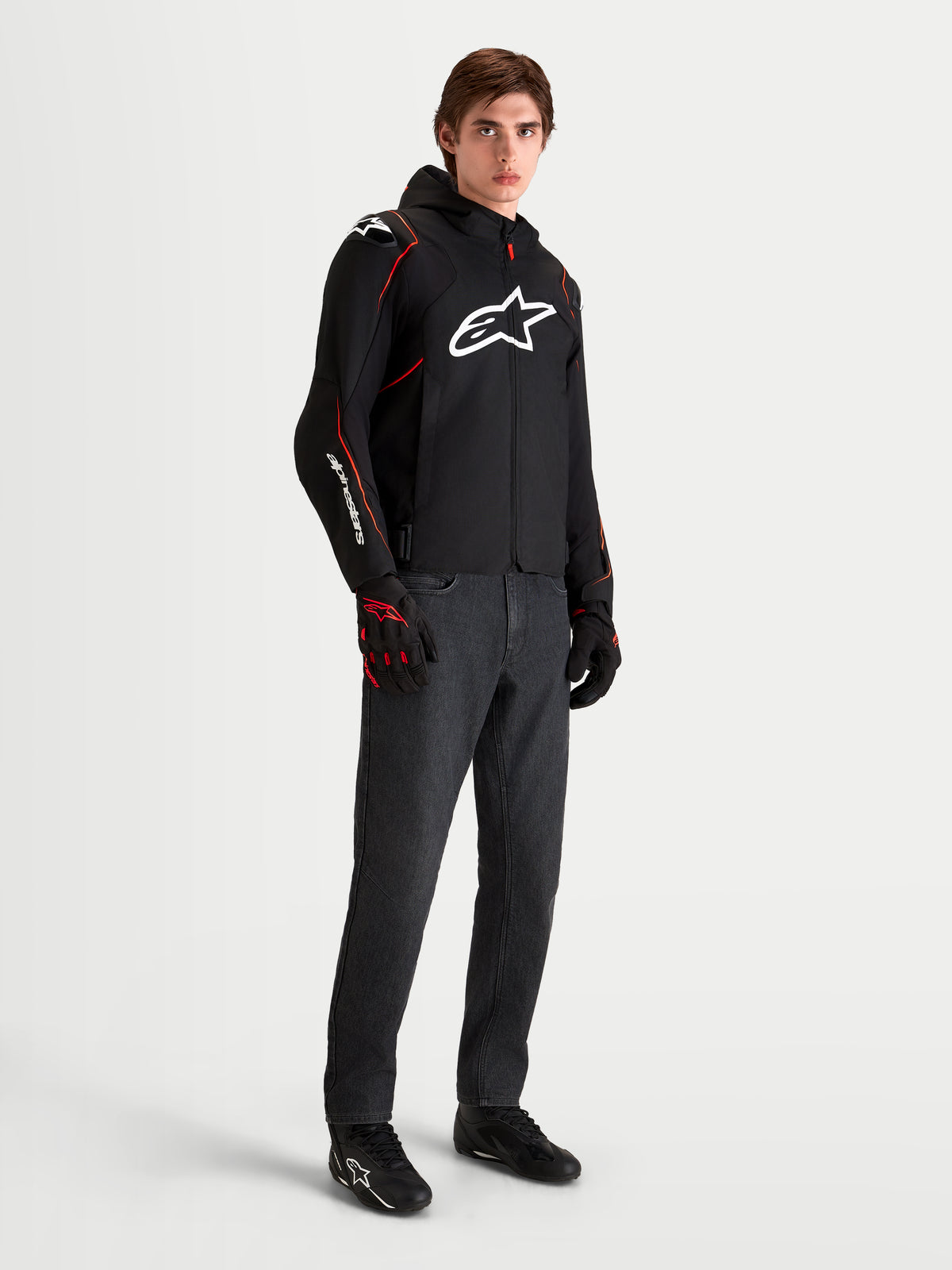 Alpinestars Zaca V2 Waterproof Giacca, giacca da moto sportiva da uomo nera e rossa con cappuccio e logo bianco sul petto, indossata da un modello con guanti da moto coordinati e jeans scuri