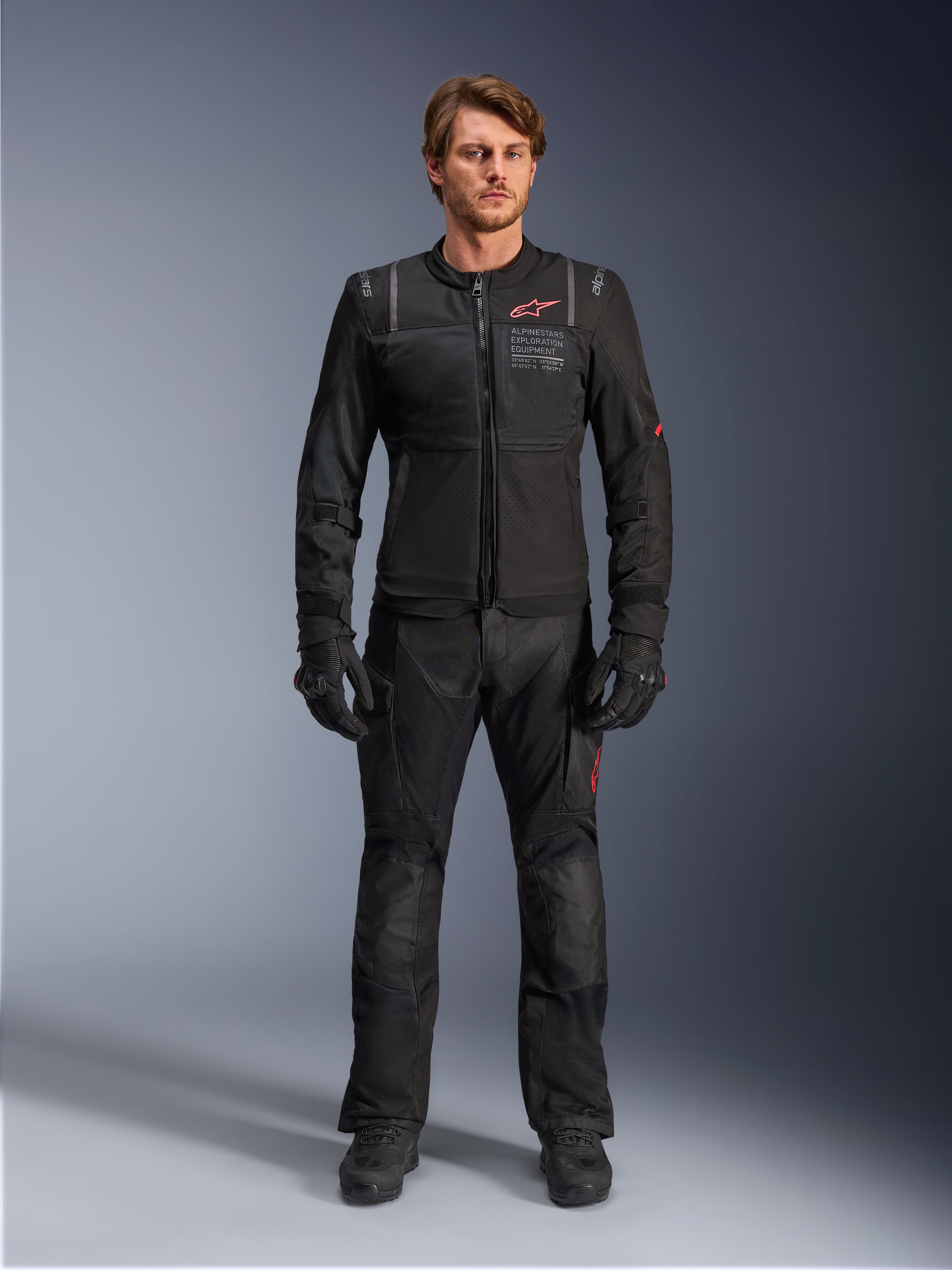 Modello con giacca Alpinestars ST-2 Air, giacca da moto nera con pannelli in mesh e logo rosso, scritta 'Alpinestars Exploration Equipment' sul petto, abbinata a pantaloni tecnici da guida e guanti neri, equipaggiamento moderno per adventure touring