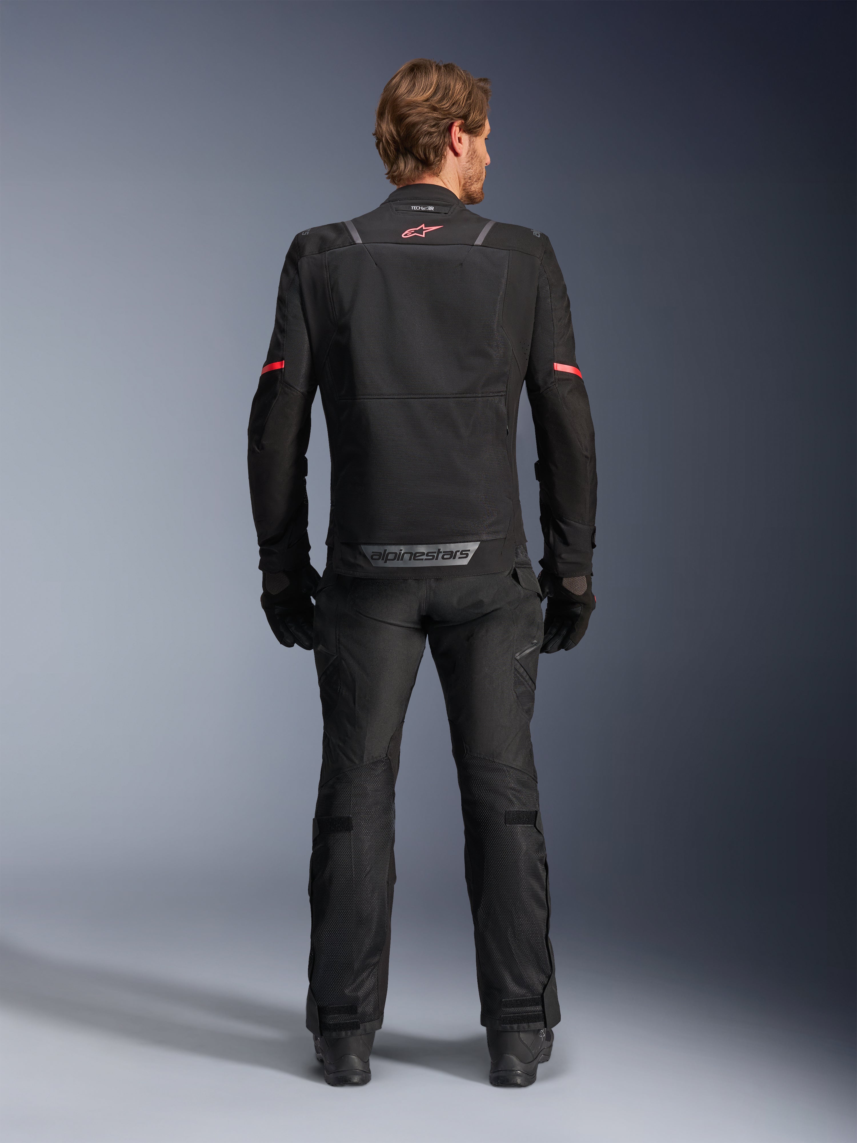 Giacca Alpinestars ST-2 Air, nero, vista posteriore di un pilota che indossa la giacca da moto e pantaloni neri abbinati, con pannelli di ventilazione in mesh, logo rosso sulla parte superiore della schiena e marchio Alpinestars grigio sul bordo inferiore posteriore