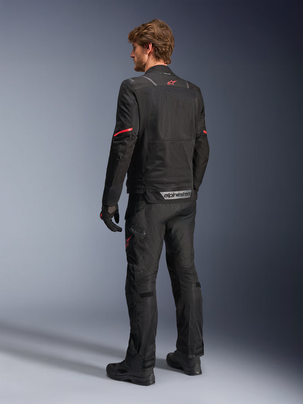 Giacca Alpinestars ST-2 Air, giacca da moto nera, vista posteriore su modello con logo a stella rosso sulla parte superiore della schiena e marchio Alpinestars riflettente in vita, abbinata a pantaloni tecnici da moto e guanti neri coordinati.