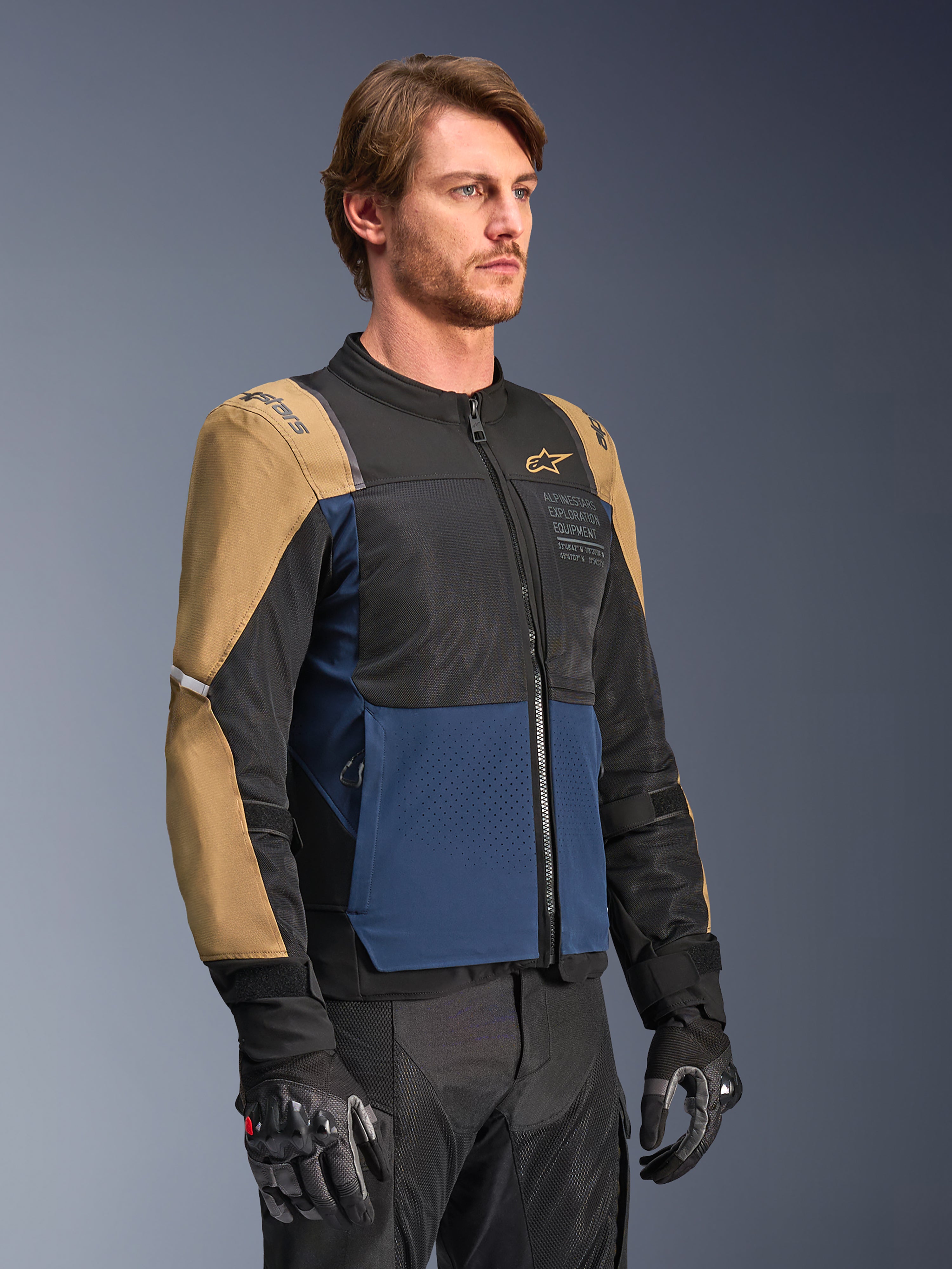 Alpinestars ST-2 Air Jacket, giacca da moto, blu profondo, kaki scuro e nero, con pannelli di ventilazione in rete ad alto flusso e marchio 'Alpinestars Exploration Equipment', indossata con guanti da moto neri, design moderno da adventure touring