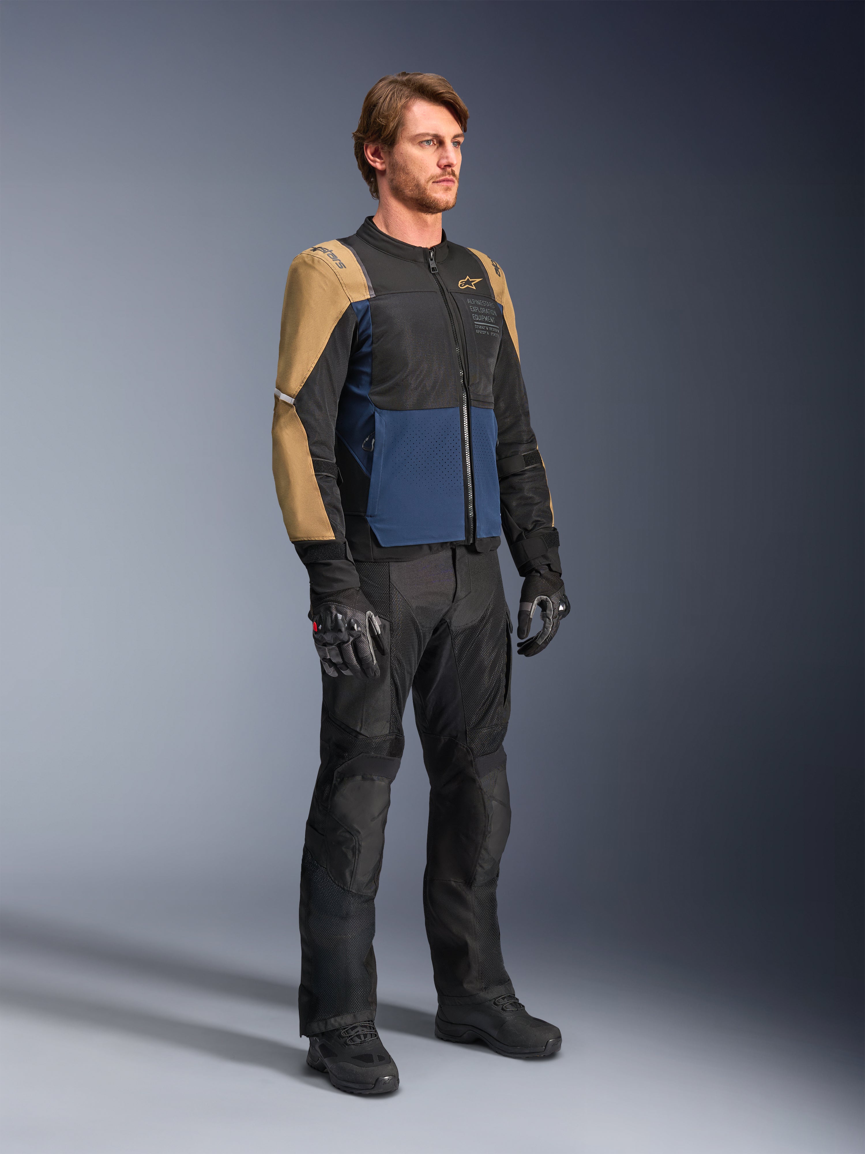Motociclista che indossa la giacca Alpinestars ST-2 Air in Deep Blue, Dark Khaki e nero, caratterizzata da ampi pannelli in rete e perforazioni laser, abbinata a pantaloni, guanti e stivali da moto neri per un look touring estivo completo.