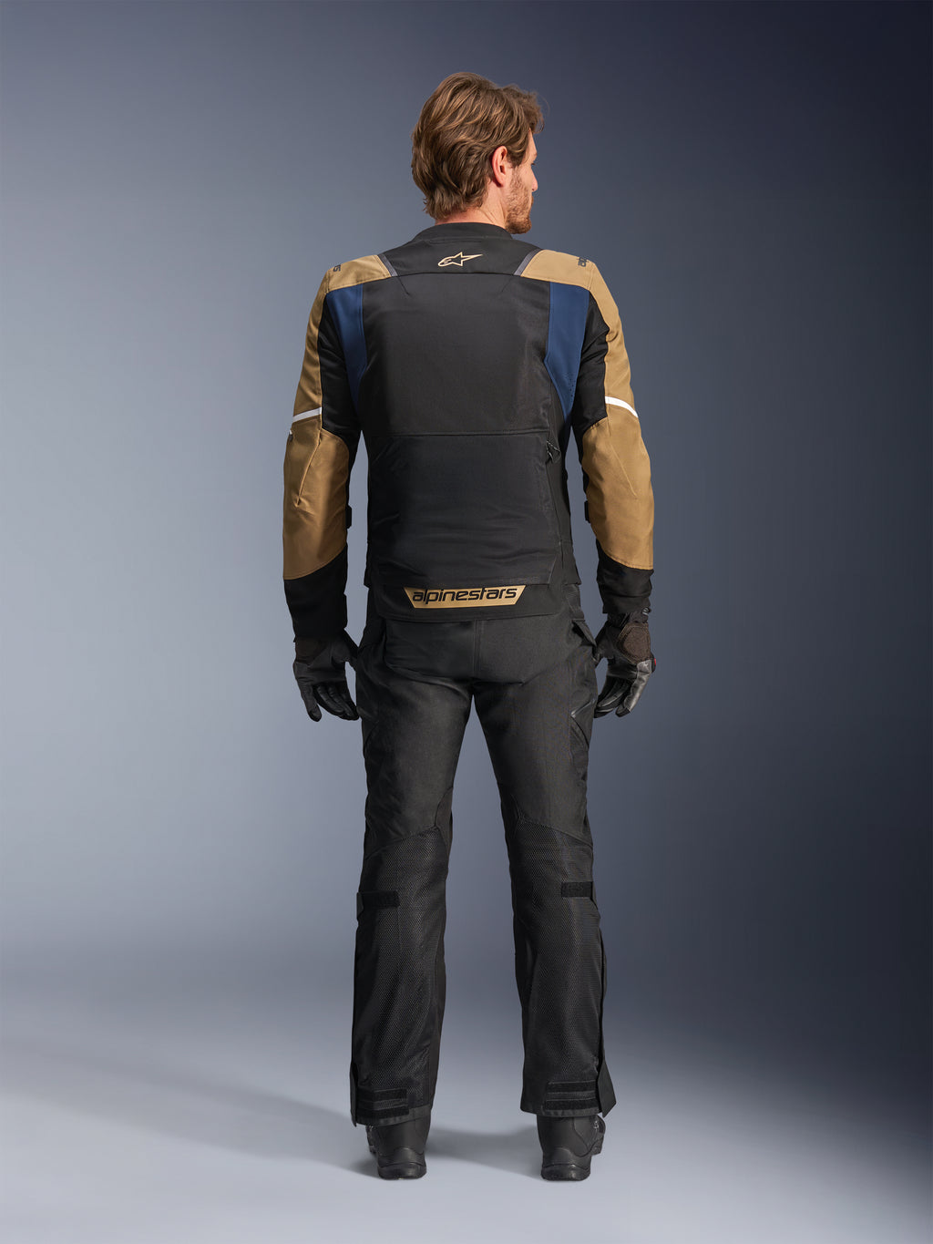 Giacca Alpinestars ST-2 Air in Deep Blue, Dark Khaki e nero, vista posteriore su modello che mostra l'ampio pannello posteriore in mesh e le maniche khaki con strisce bianche, abbinata a pantaloni tecnici da moto e guanti neri per l'adventure touring.