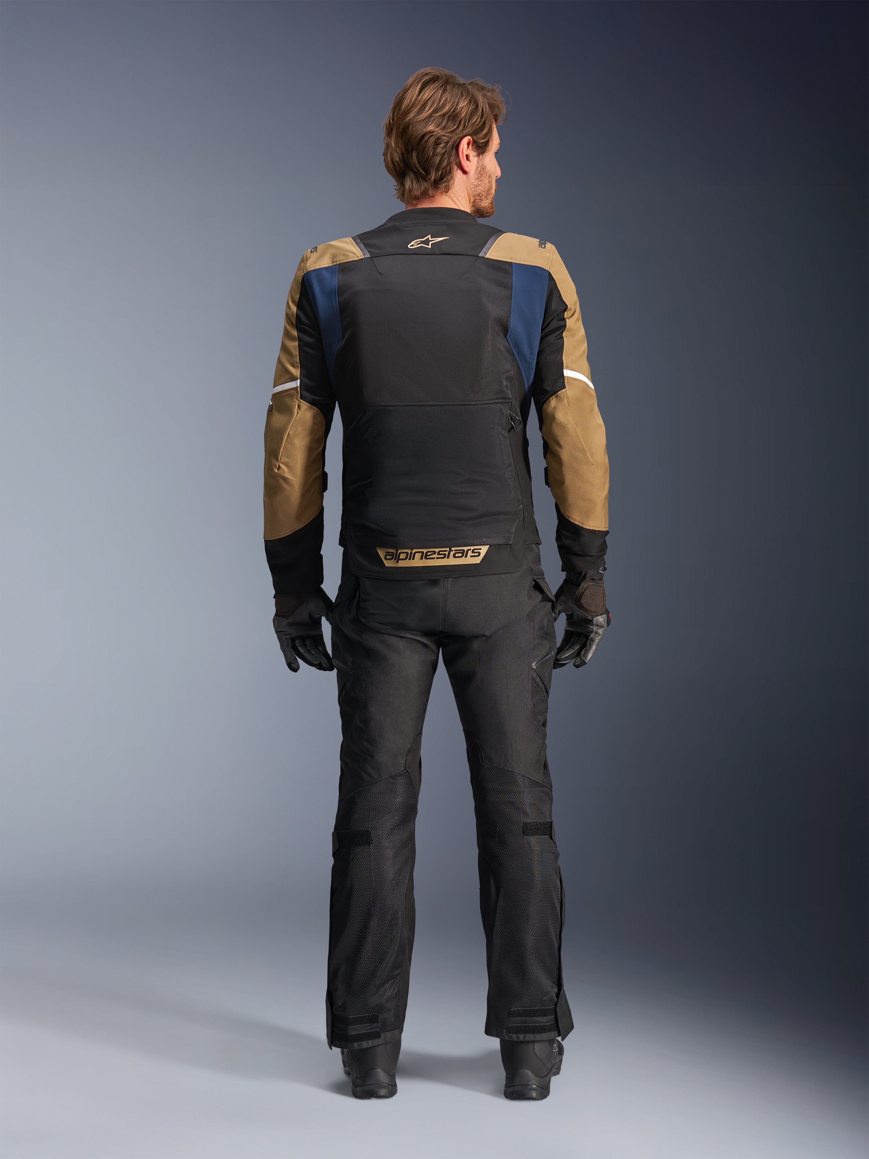 Giacca Alpinestars ST-2 Air in Deep Blue, Dark Khaki e nero, vista posteriore su modello che mostra l'ampio pannello posteriore in mesh e le maniche khaki con strisce bianche, abbinata a pantaloni tecnici da moto e guanti neri per l'adventure touring.