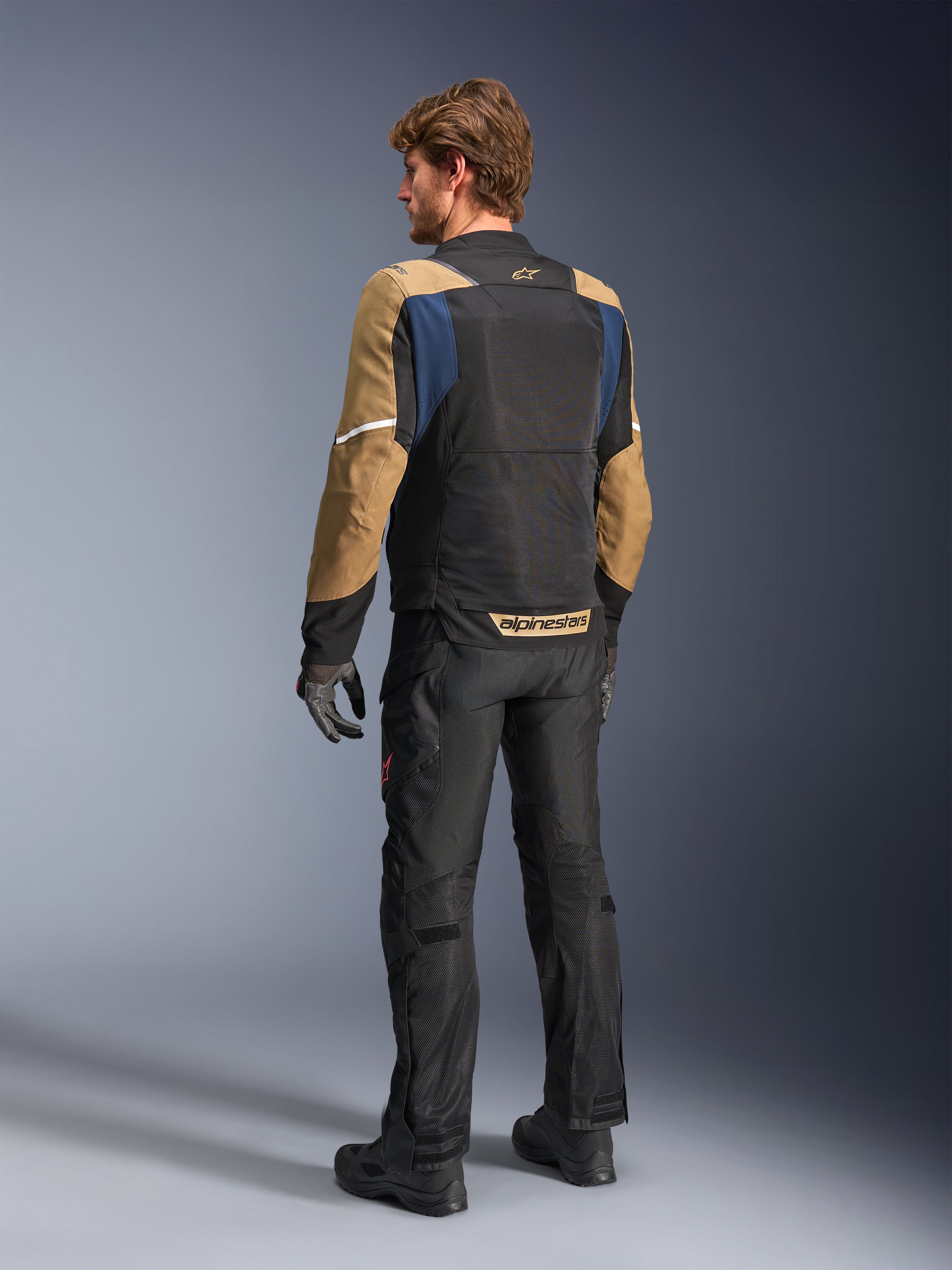 Alpinestars ST-2 Air Jacket, giacca da moto touring estiva, blu scuro, kaki scuro e nero, vista posteriore su modello che mostra pannelli in rete, maniche kaki e accenti blu, con strisce riflettenti bianche e marchio evidente sulla parte bassa della schiena, abbinata a pantaloni da moto neri.
