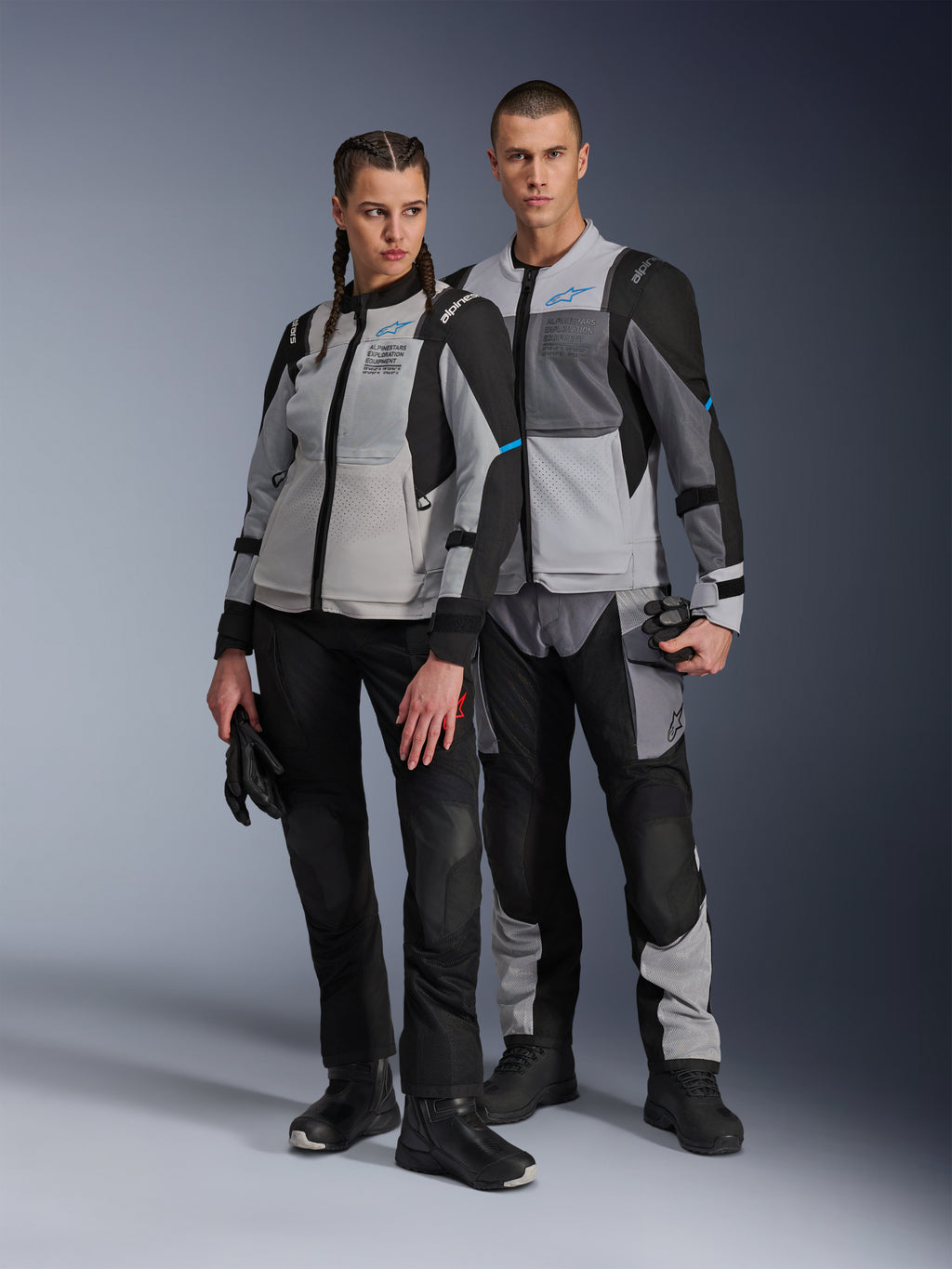 Uomo e donna con giacche Alpinestars Stella ST-2 Air e pantaloni tecnici coordinati, colorazione Ice Gray e Black con accenti blu, caratterizzati da ventilazione in mesh e marchio 'ALPINESTARS EXPLORATION EQUIPMENT', scatto di coppia in studio