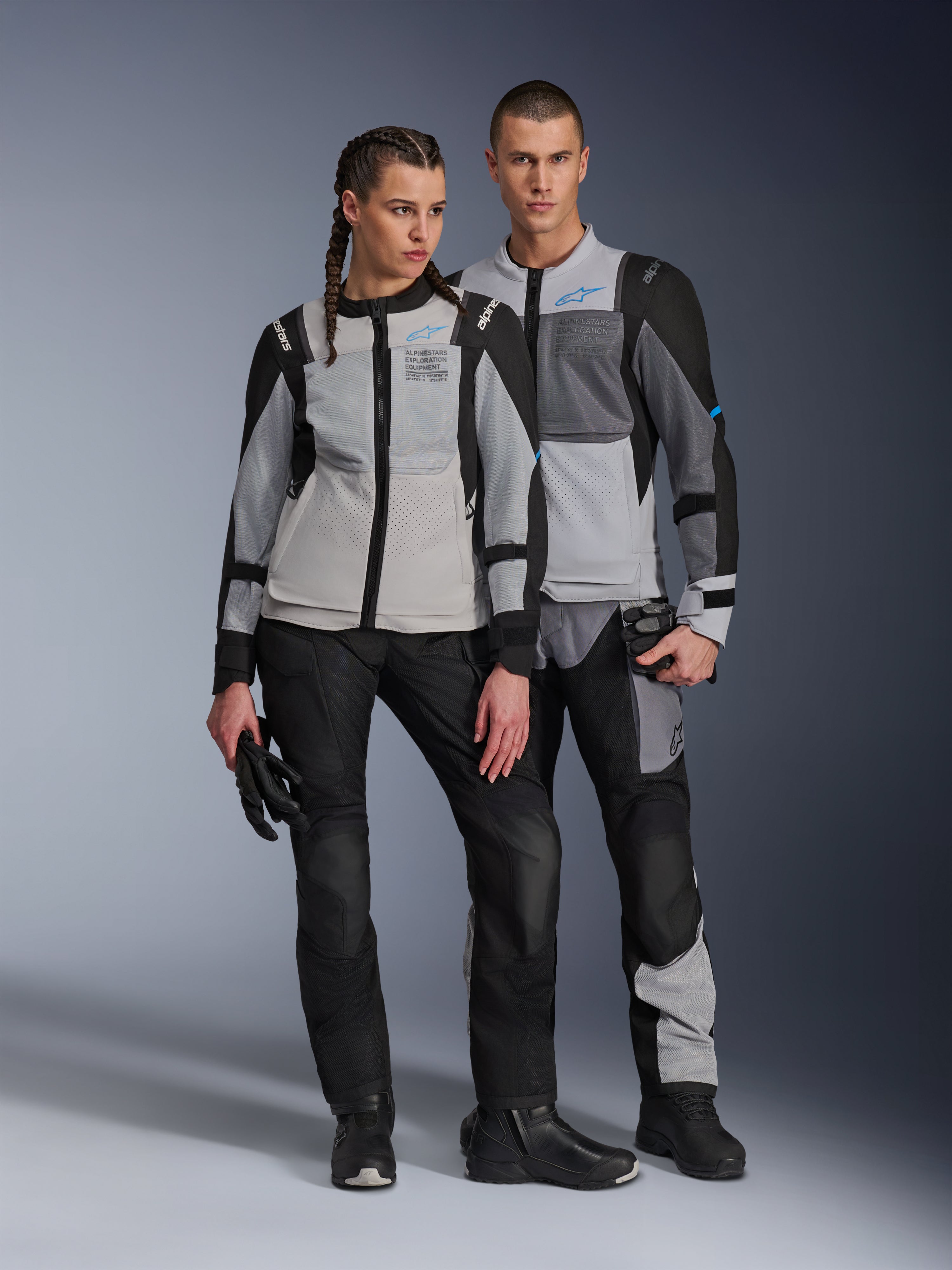 Modelli uomo e donna che indossano giacche Alpinestars ST-2 Air in Ice Gray, Dark Gray e Nero, abbigliamento tecnico da mototurismo estivo con ampi pannelli in rete e branding Exploration Equipment, abbinati a pantaloni, guanti e stivali da moto neri.