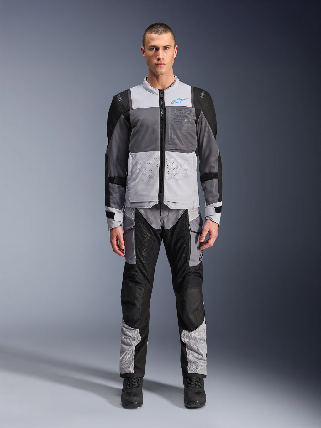 Giacca Alpinestars ST-2 Air, giacca da moto touring, grigio ghiaccio, grigio scuro e nero, indossata da un modello con pantaloni adventure abbinati, con ampi pannelli in rete, logo Alpinestars blu sul petto e marchio Alpinestars Exploration Equipment.