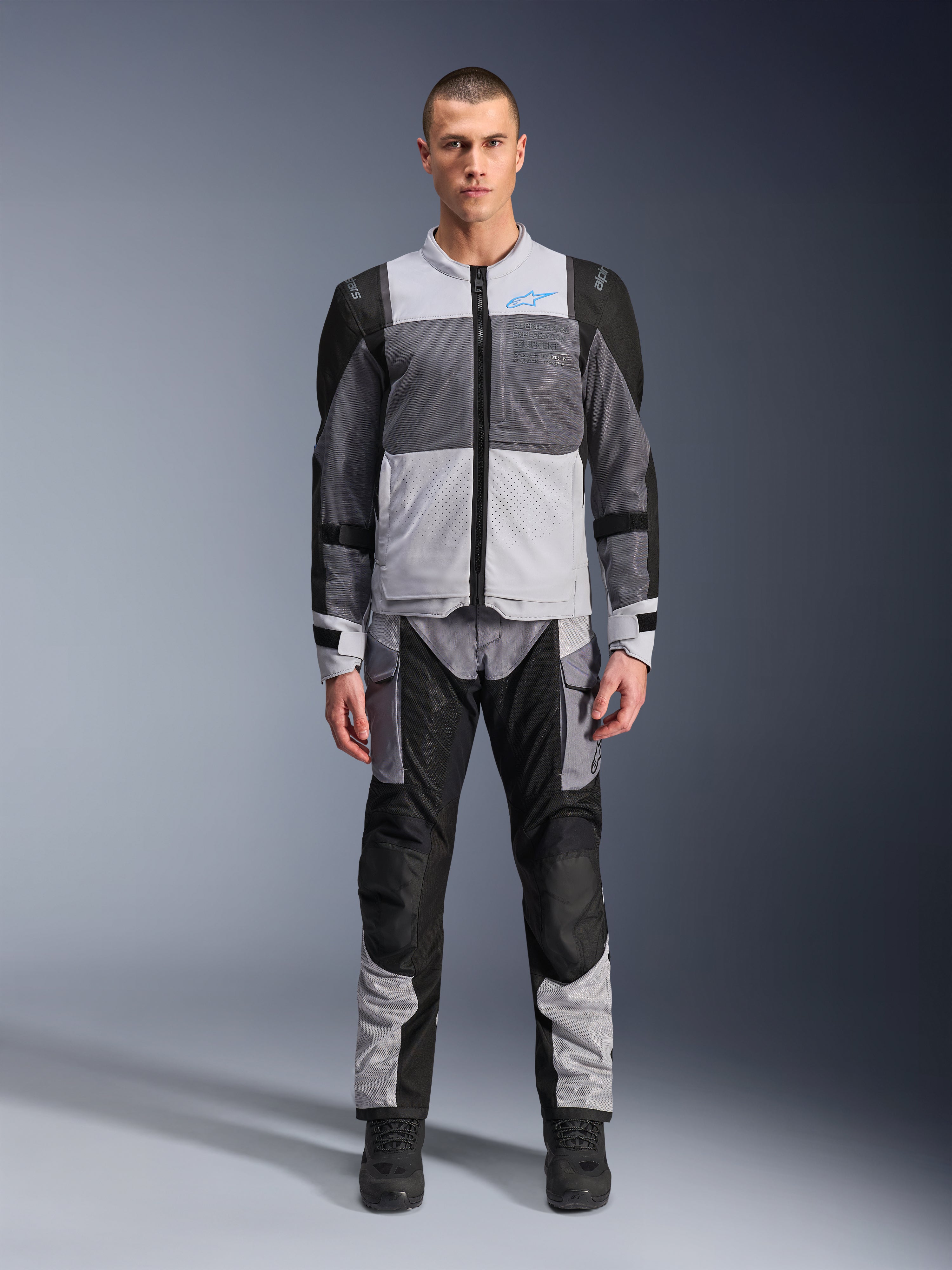 Giacca Alpinestars ST-2 Air, giacca da moto touring, grigio ghiaccio, grigio scuro e nero, indossata da un modello con pantaloni adventure abbinati, con ampi pannelli in rete, logo Alpinestars blu sul petto e marchio Alpinestars Exploration Equipment.