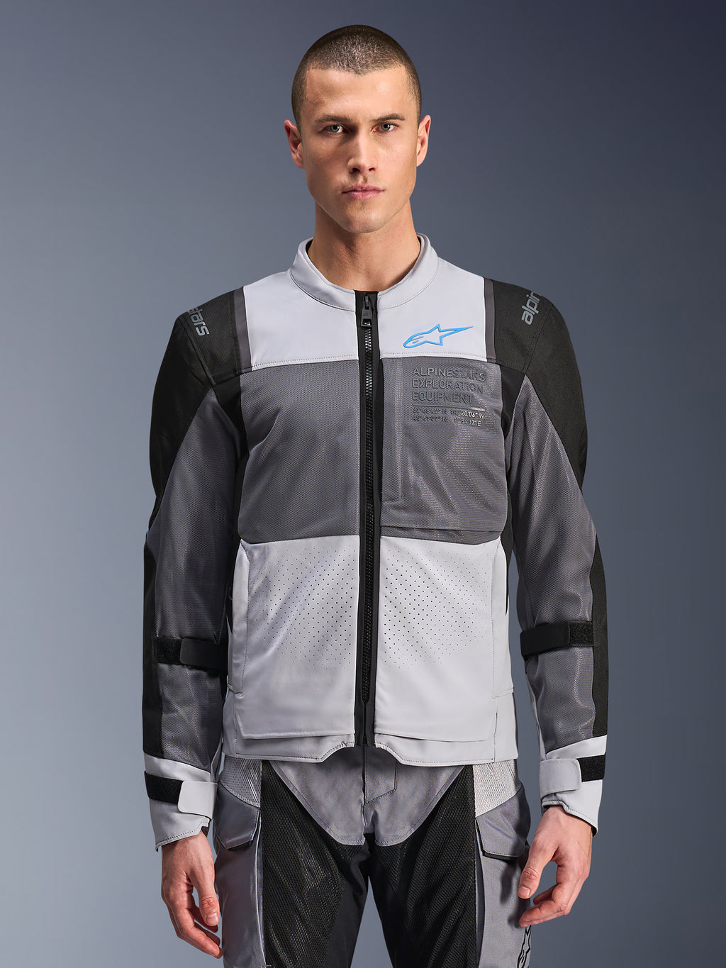 Giacca Alpinestars ST-2 Air, giacca da moto da uomo, grigio ghiaccio e grigio scuro con accenti neri, ampi pannelli di ventilazione in rete, parte anteriore traforata al laser, logo Alpinestars blu, cerniera frontale, indossata da un modello con pantaloni da moto coordinati