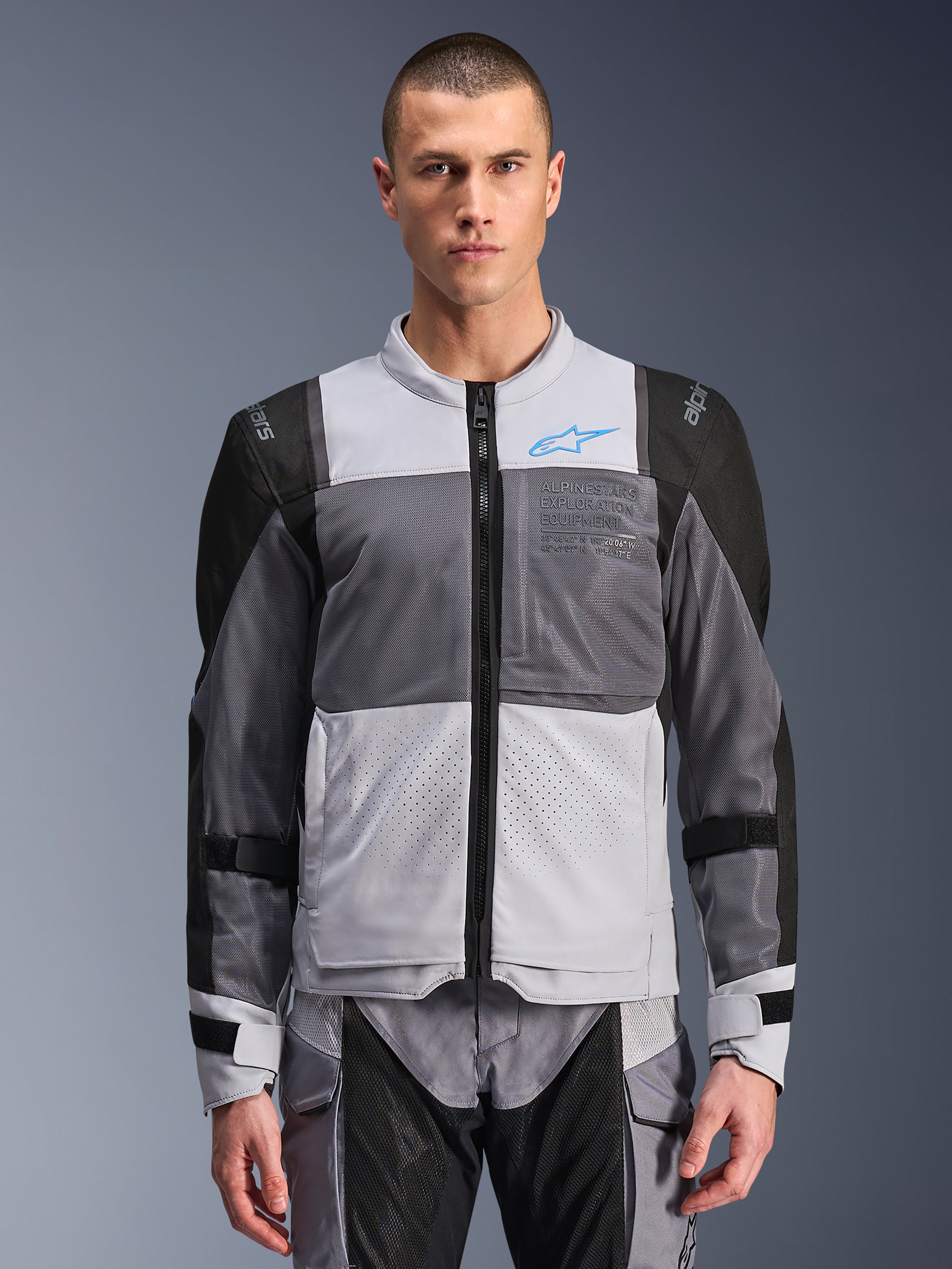 Giacca Alpinestars ST-2 Air, giacca da moto da uomo, grigio ghiaccio e grigio scuro con accenti neri, ampi pannelli di ventilazione in rete, parte anteriore traforata al laser, logo Alpinestars blu, cerniera frontale, indossata da un modello con pantaloni da moto coordinati