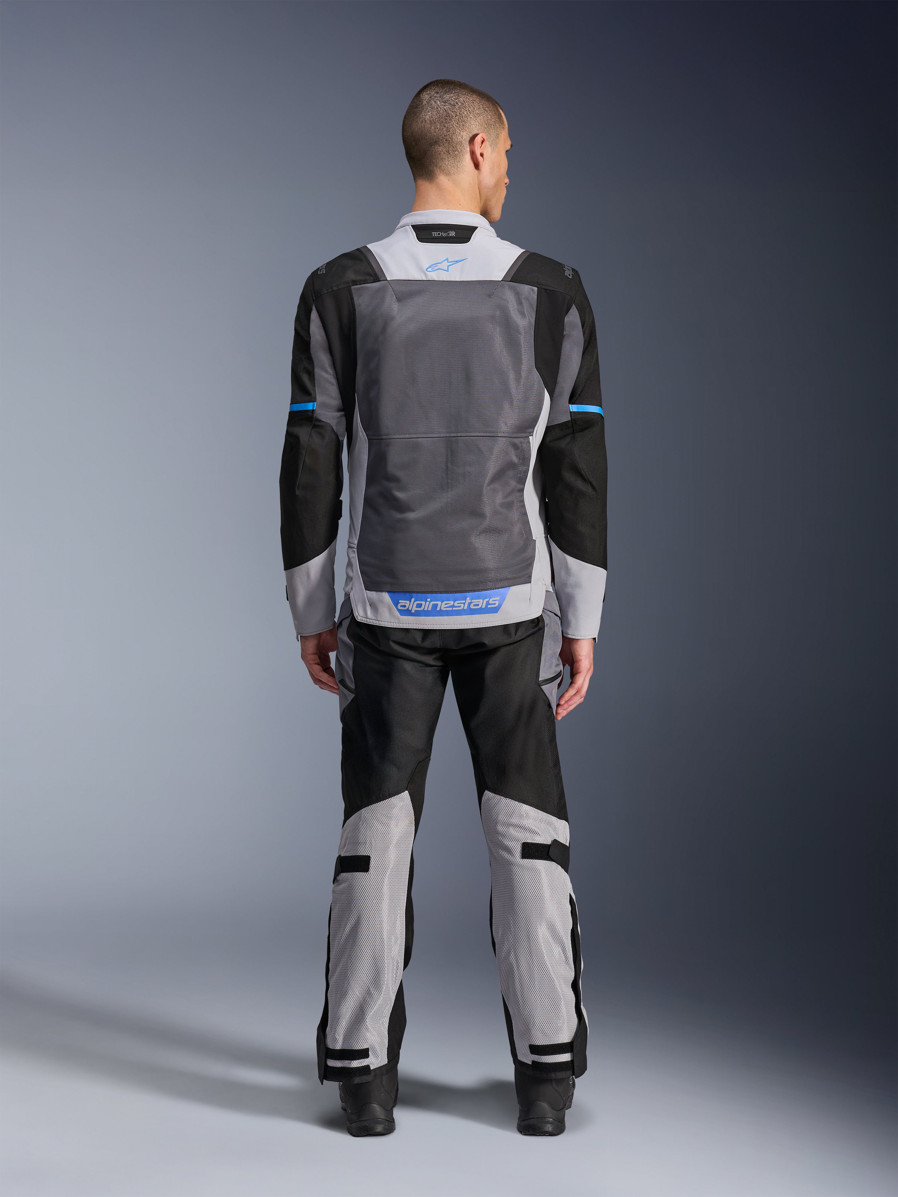 Giacca Alpinestars ST-2 Air, Ice Gray Dark Gray Black, vista posteriore su modello, con ampi pannelli di ventilazione in rete, dettagli blu e logo Alpinestars, abbinata a pantaloni da moto coordinati per adventure touring.