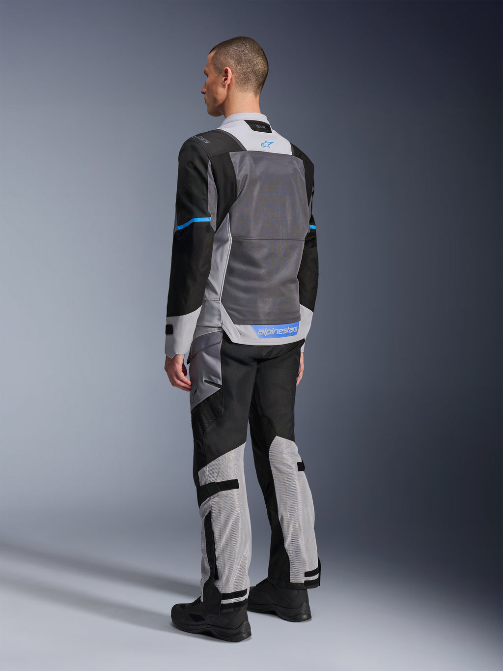 Modello maschile visto da dietro che indossa la giacca Alpinestars ST-2 Air in Ice Gray, Dark Gray e nero, con un ampio pannello posteriore in rete grigia, dettagli del logo blu e pantaloni da moto coordinati per il touring estivo.