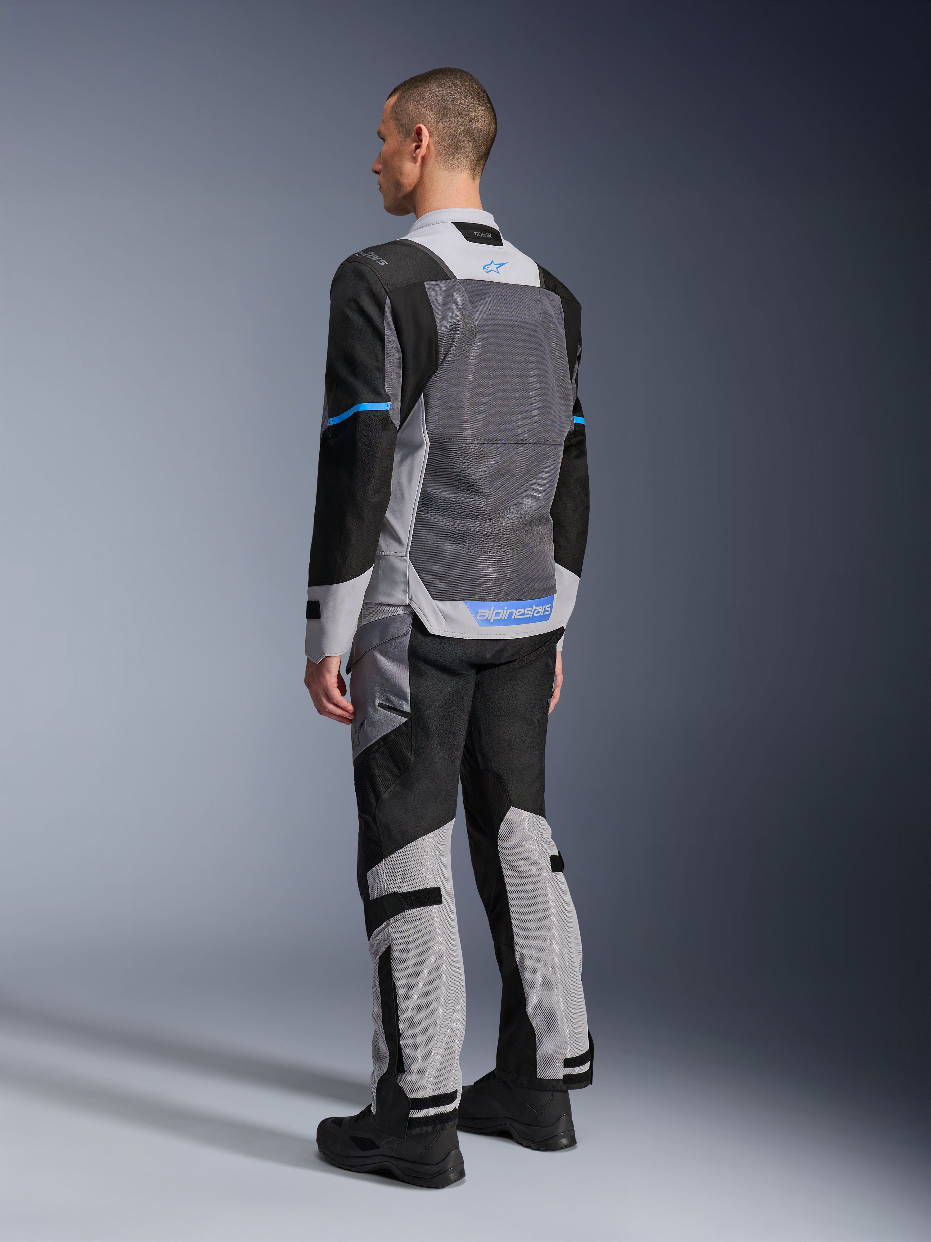 Modello maschile visto da dietro che indossa la giacca Alpinestars ST-2 Air in Ice Gray, Dark Gray e nero, con un ampio pannello posteriore in rete grigia, dettagli del logo blu e pantaloni da moto coordinati per il touring estivo.
