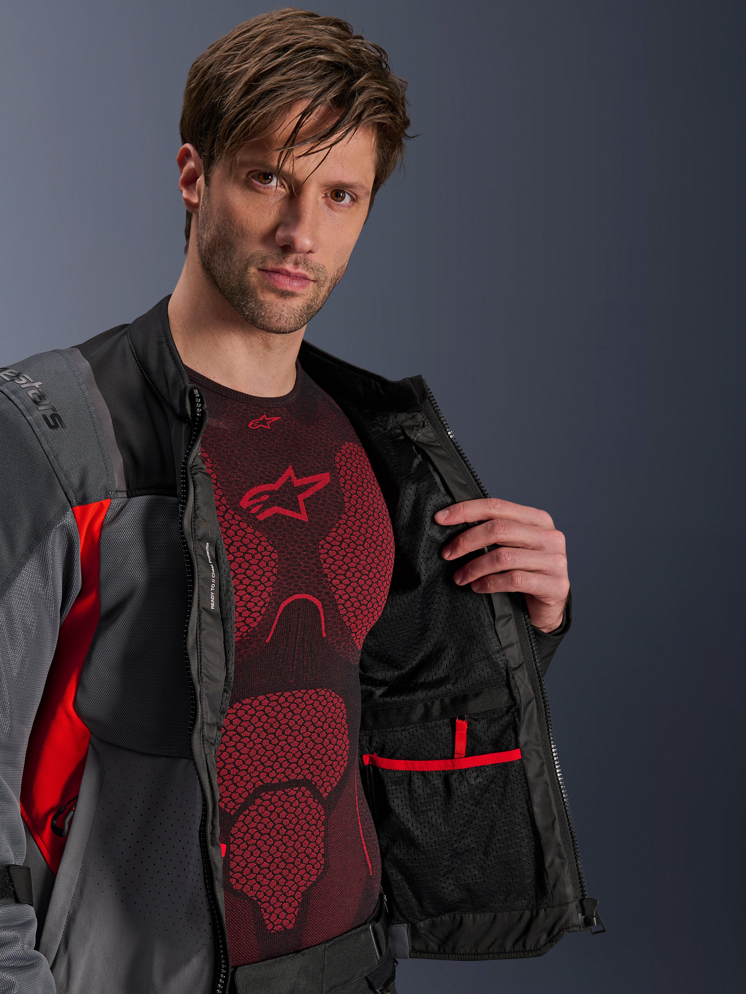 Giacca Alpinestars ST-2 Air, grigio scuro e rosso acceso, aperta per mostrare la fodera interna in rete e la tasca con profili rossi, indossata sopra un base layer tecnico rosso, abbigliamento moderno da moto touring adventure