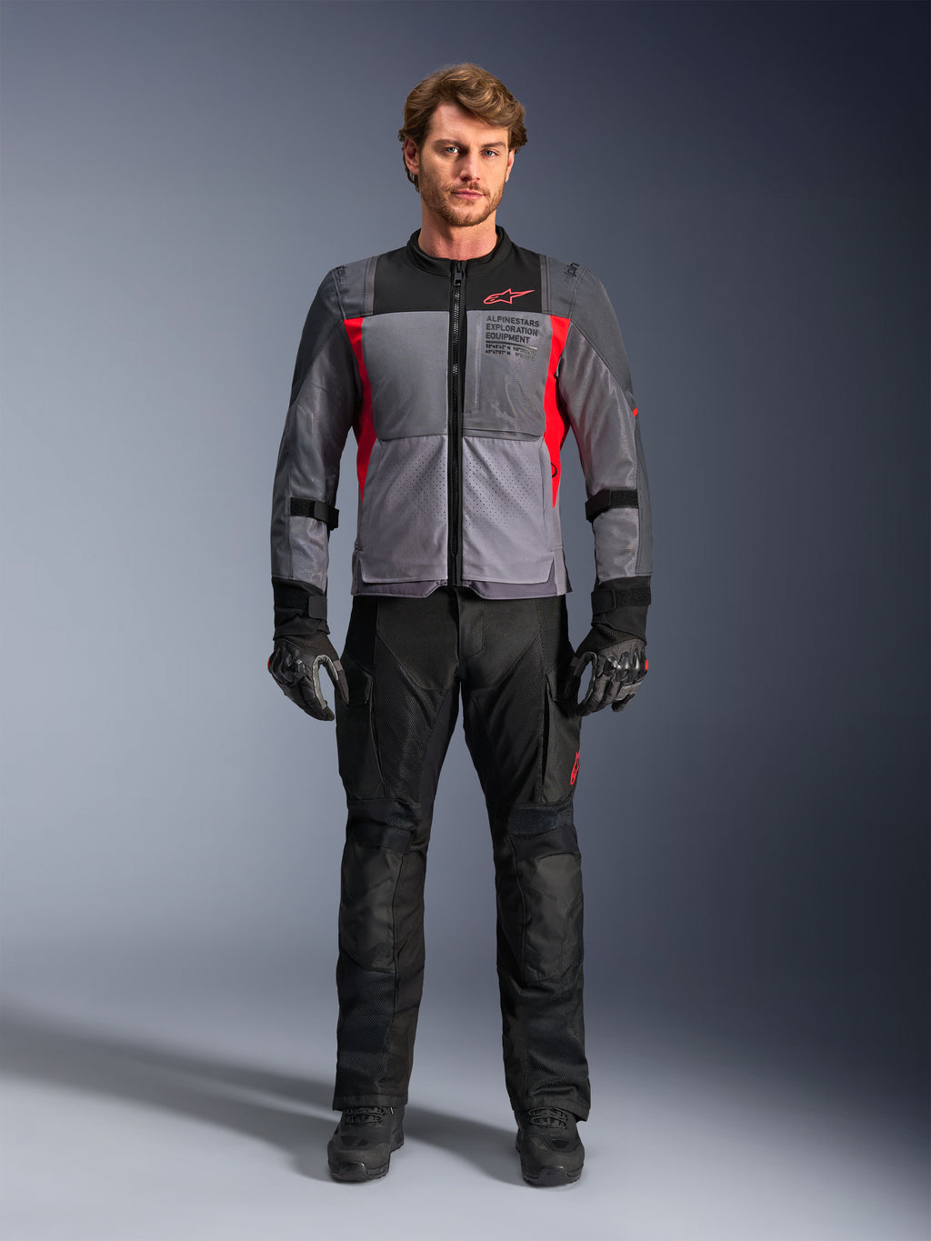Giacca Alpinestars ST-2 Air in grigio scuro e rosso acceso, indossata da un modello con pantaloni e guanti da moto neri, dotata di ampi pannelli in rete per la ventilazione e marchio moderno exploration equipment