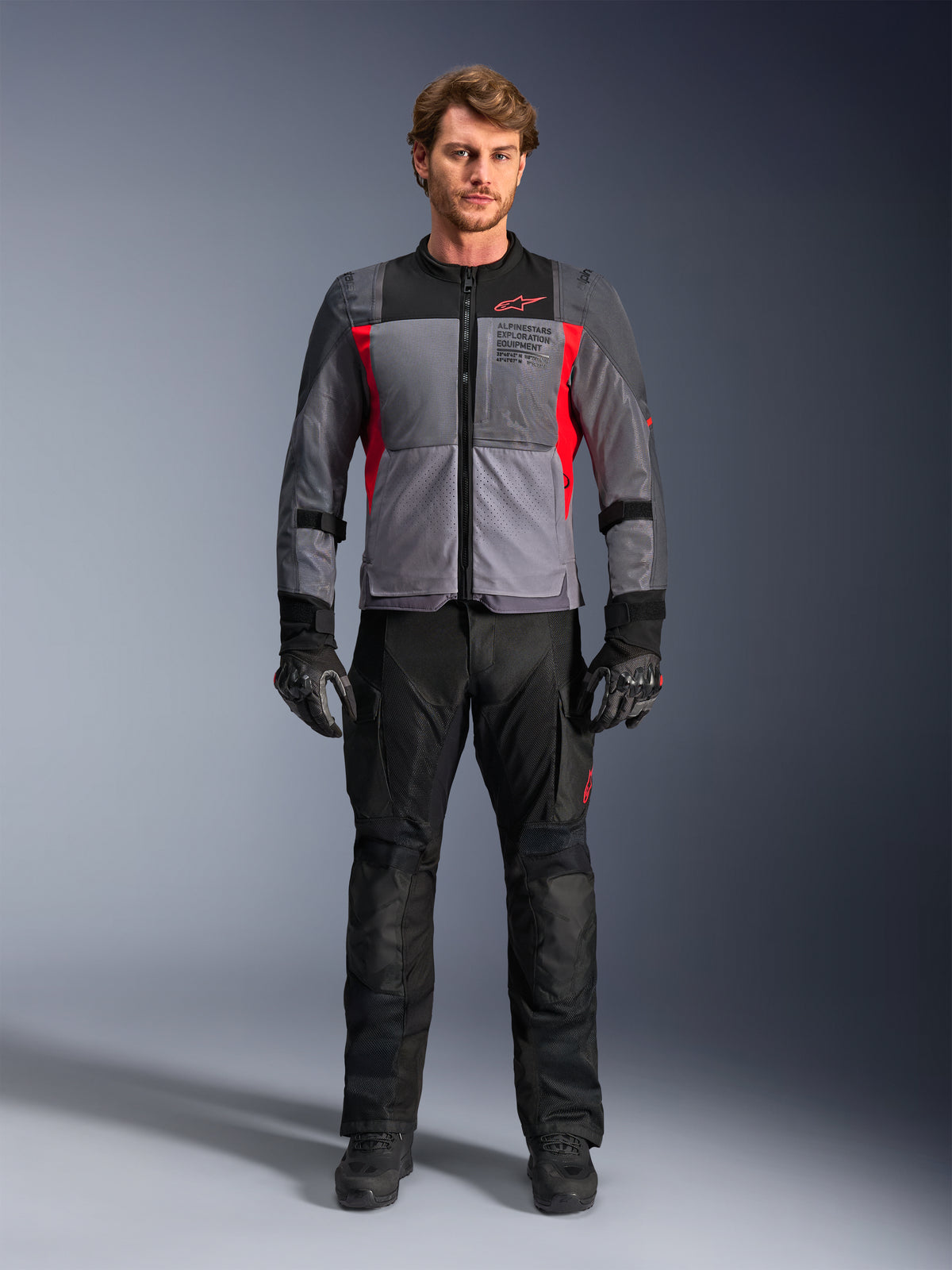 Giacca Alpinestars ST-2 Air in grigio scuro e rosso acceso, indossata da un modello con pantaloni e guanti da moto neri, dotata di ampi pannelli in rete per la ventilazione e marchio moderno exploration equipment