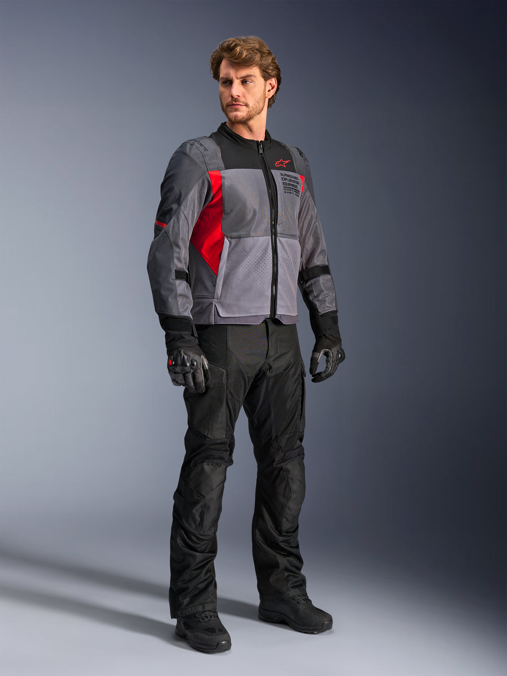 Modello maschile che indossa la giacca Alpinestars ST-2 Air, giacca da moto in tessuto, grigio scuro e rosso brillante, abbinata a pantaloni, guanti e stivali neri, con ampi pannelli in rete, marchio Alpinestars Exploration Equipment e un'estetica touring moderna